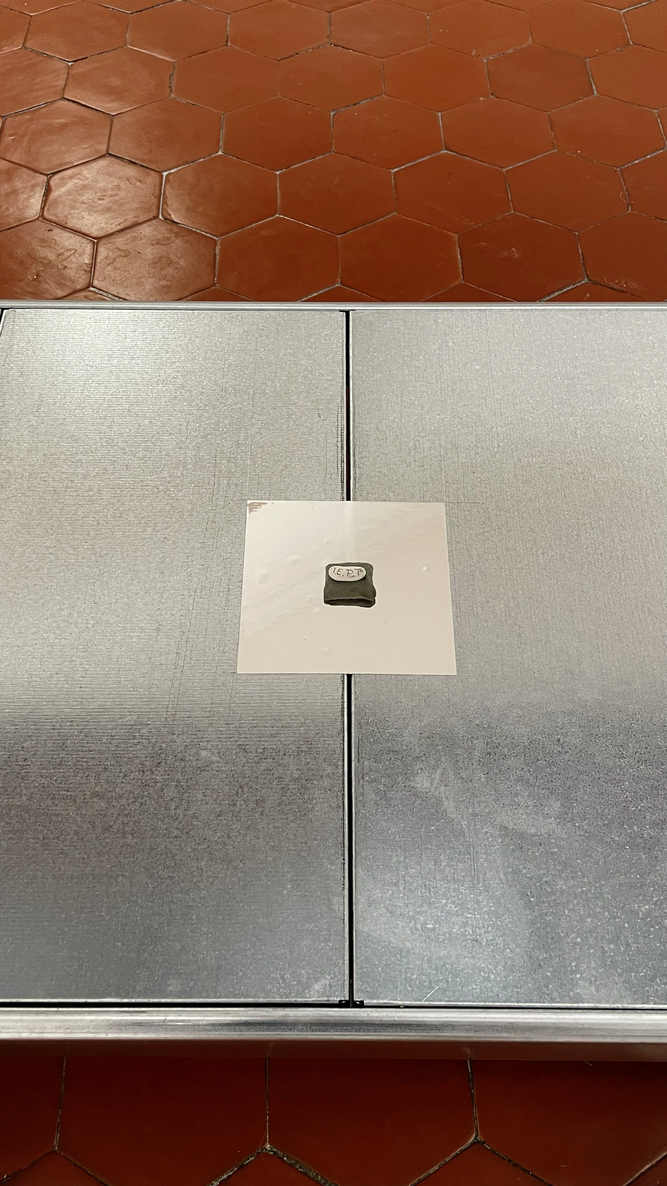 A metal table with a small black object on it, placed on a white paper, with red hexagonal tile flooring visible underneath.