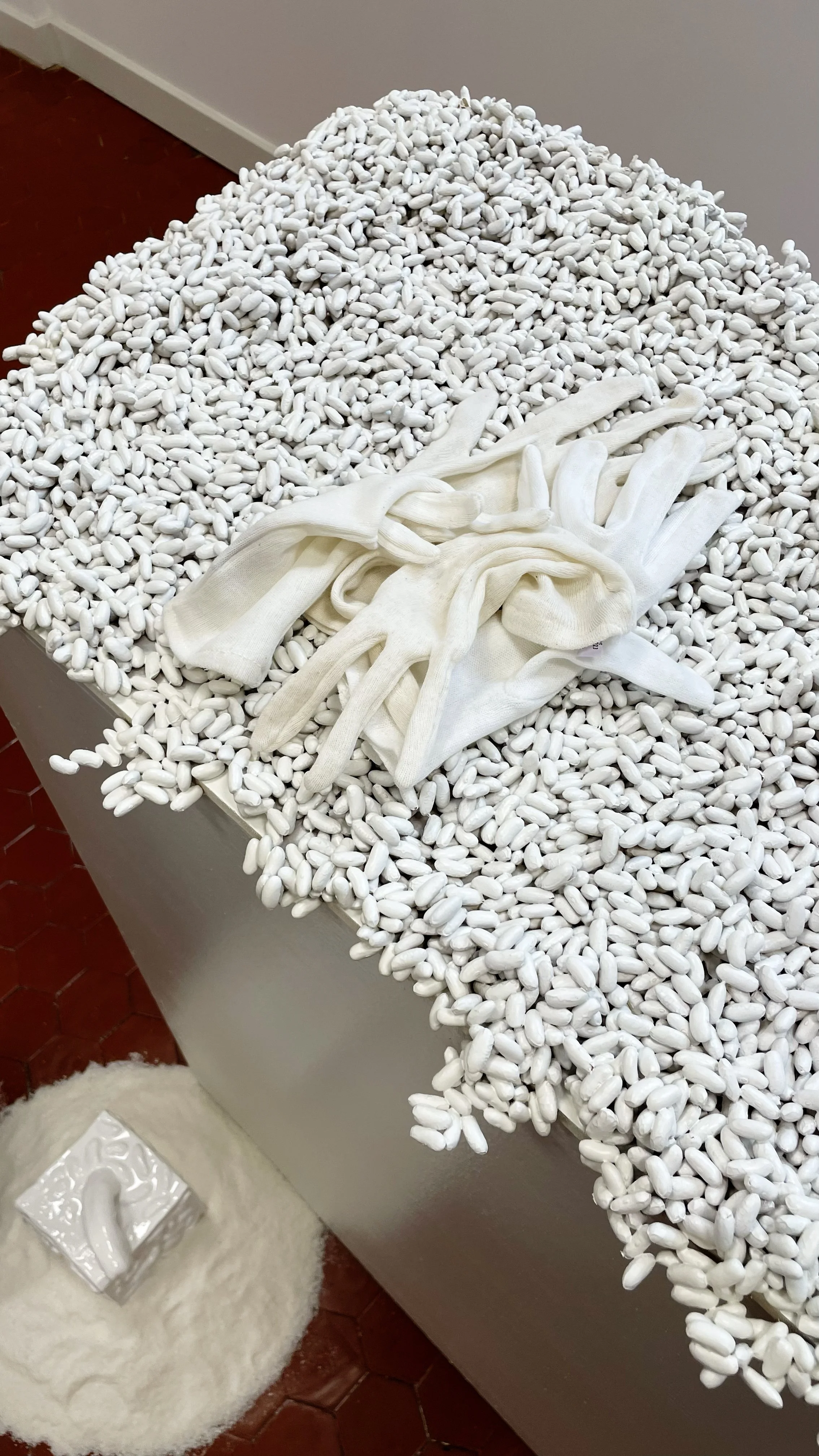 A display of white ceramic or porcelain objects resembling beans, with a pair of white gloves placed on top. A fluffy stool with a tissue box resting on it is positioned nearby.