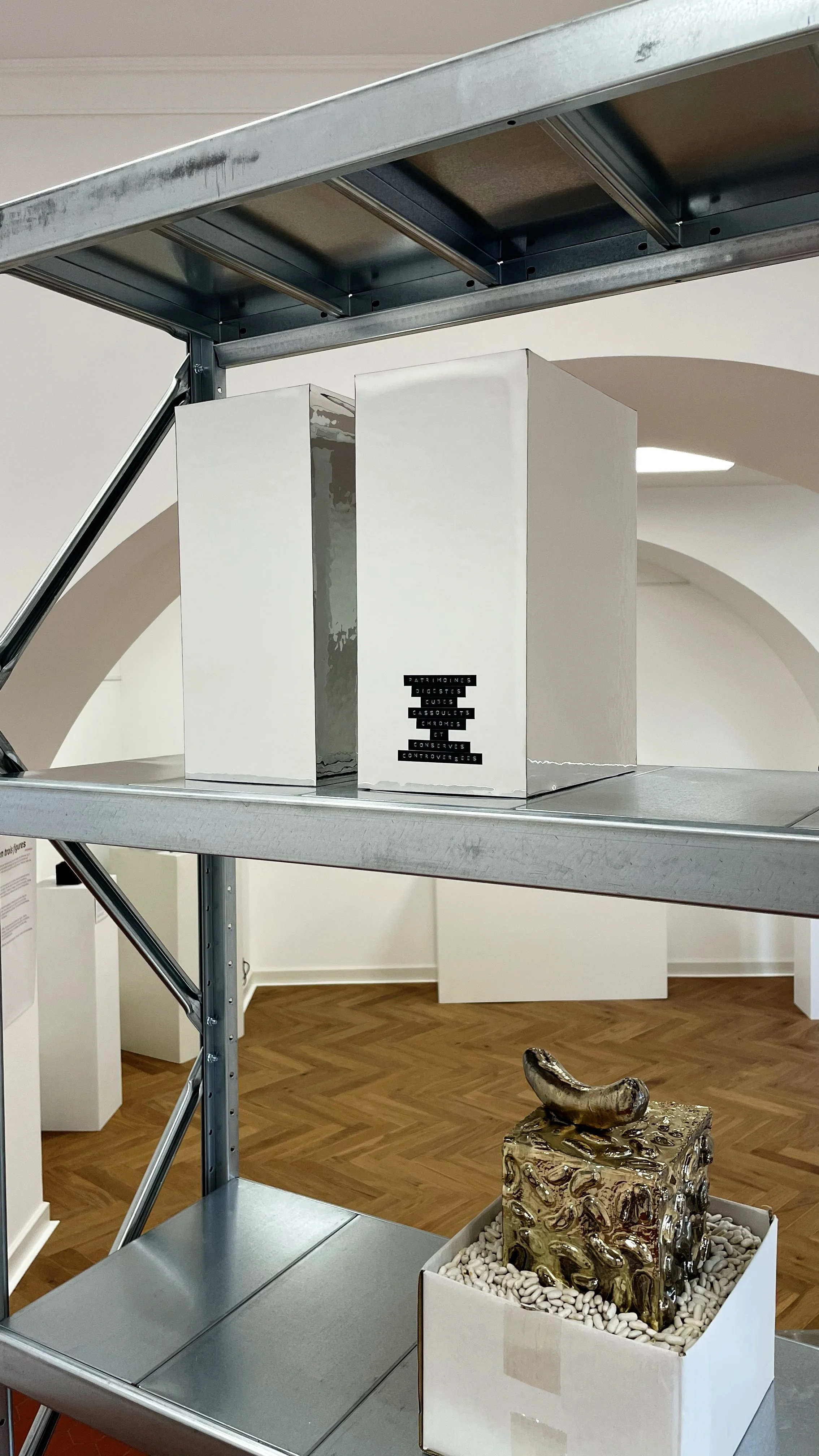 Art exhibit showing a metallic sculpture of a shoe or boot on a textured block with pebbles around it, and a white box with black labels and text.