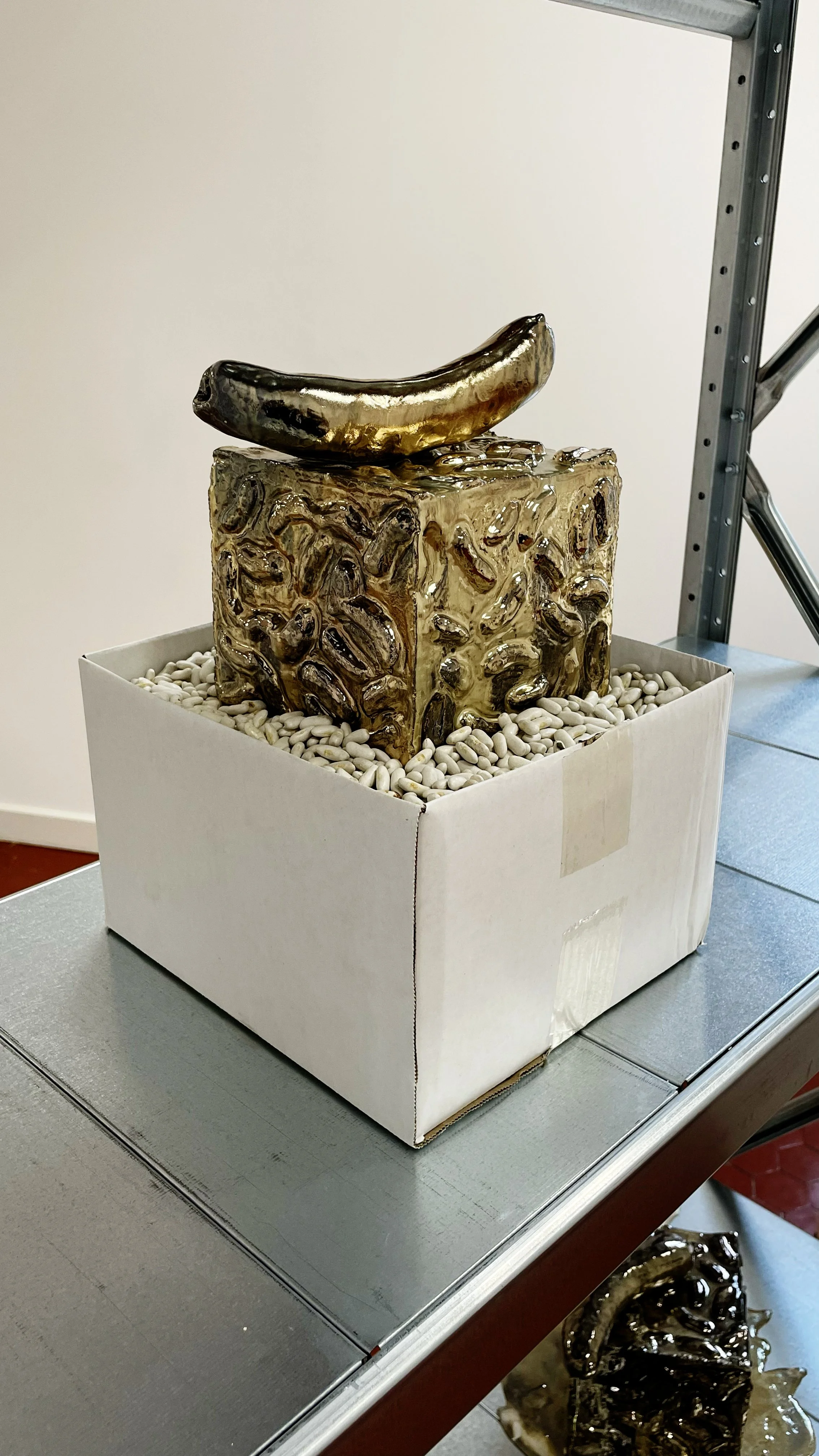 Ceramic sculpture of a banana on top of a textured block, displayed in a white box filled with white pebbles, placed on a metallic table.
