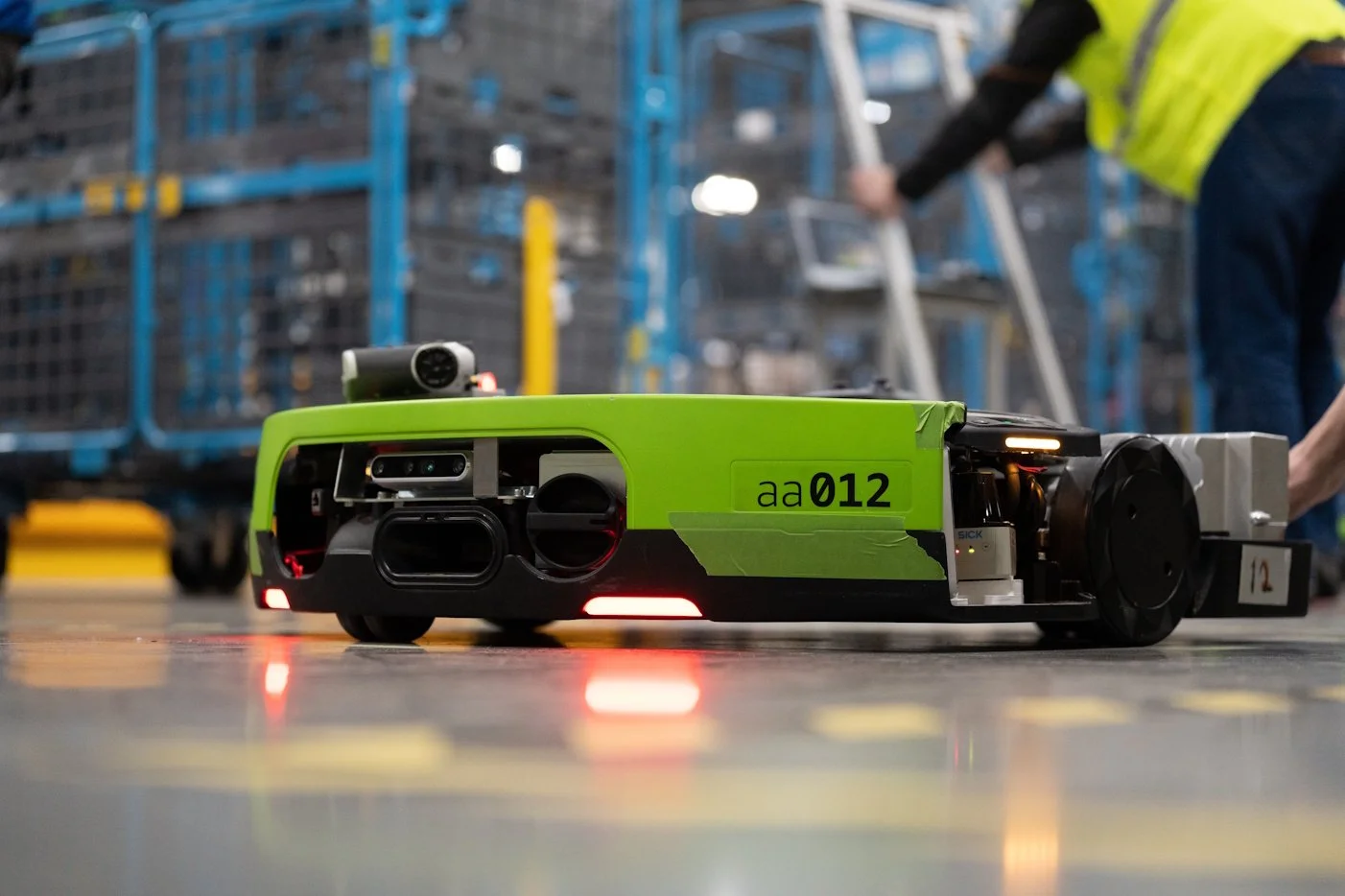 A green autonomous robot on the floor of an industrial or warehouse setting, with a person in safety clothing near scaffolding or shelving in the background.