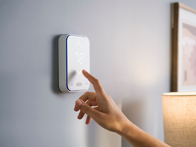 A person presses a touch screen thermostat on a wall, displaying the number 72.