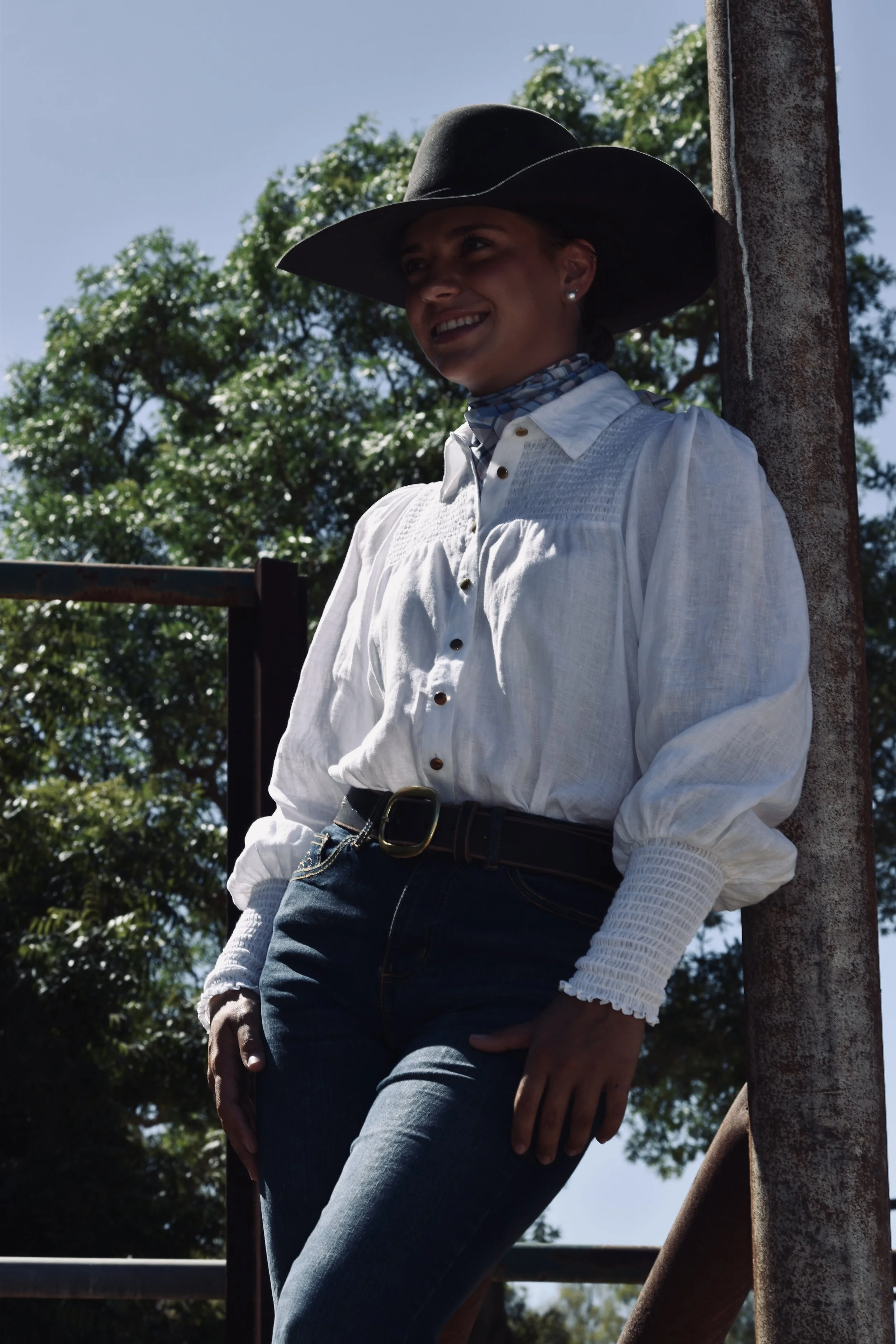 A woman wearing a black wide-brimmed hat, a white long-sleeve blouse, and dark jeans, leaning against a rusty metal pole outdoors with green trees and a cloudy sky in the background.
