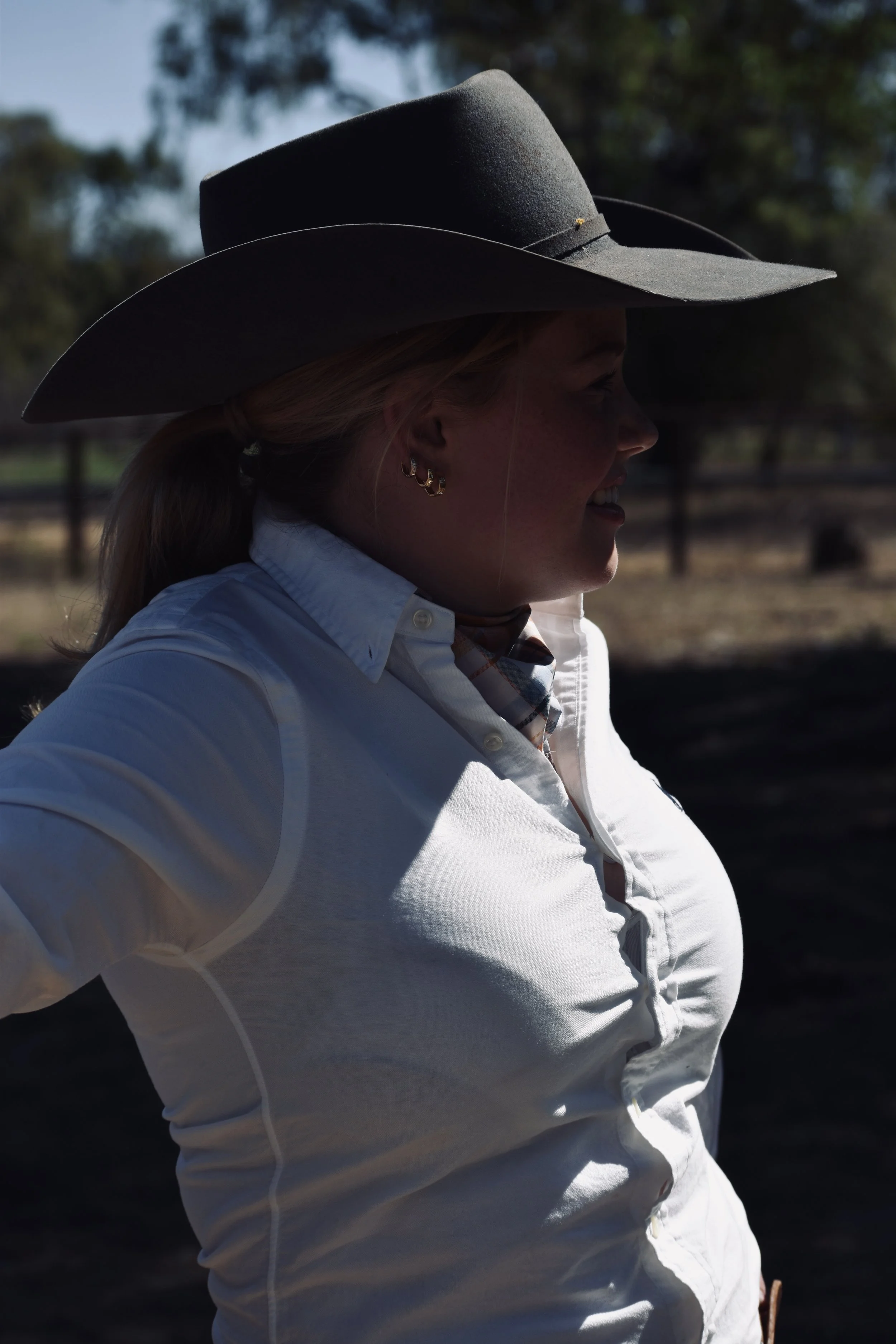 A woman wearing a large gray cowboy hat, a white button-up shirt, and hoop earrings, standing outdoors with trees and a fence in the background.