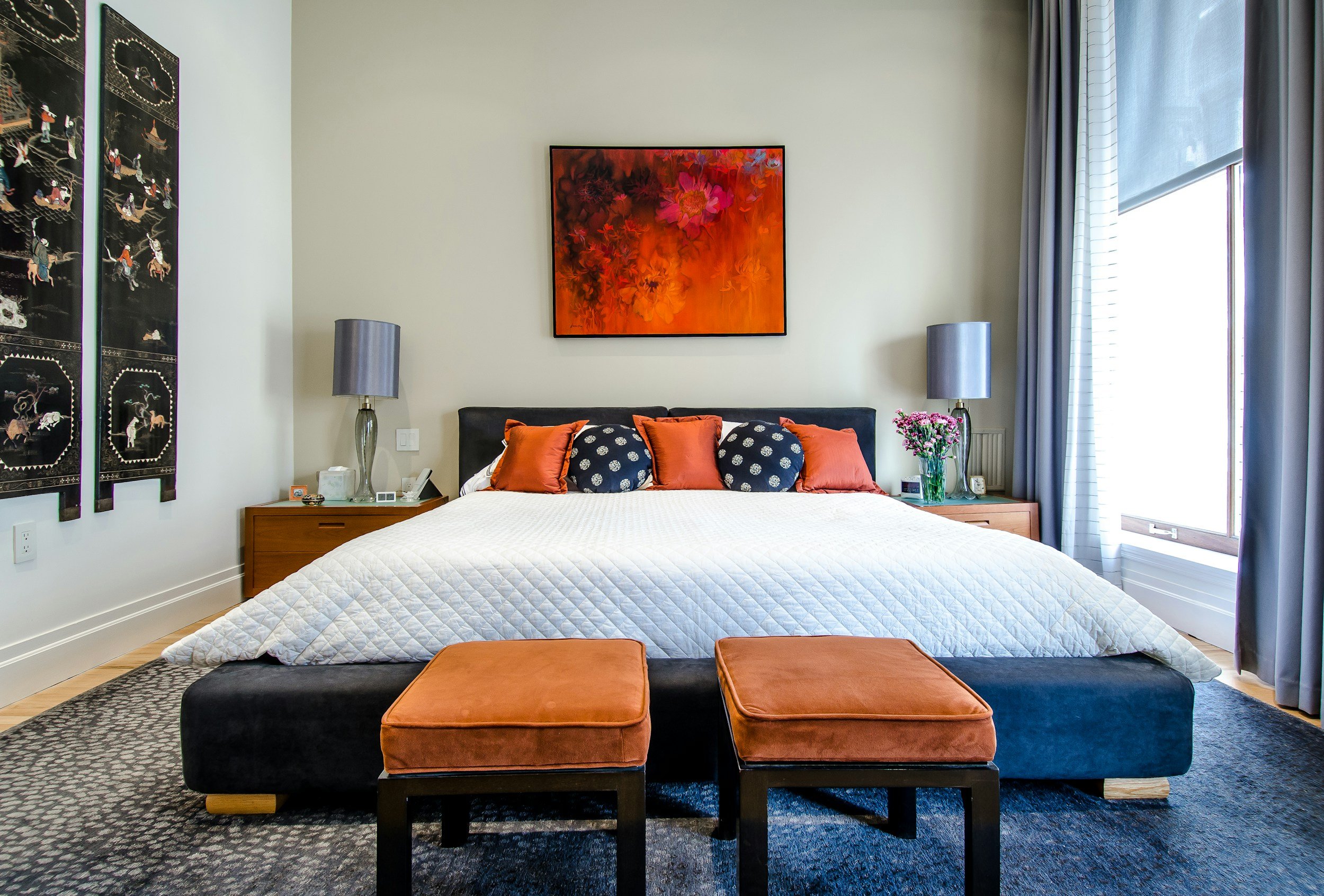 Ein modernes Schlafzimmer mit einem großen Bett, weißen Bettdecke, mehreren orangefarbenen und schwarzen Kissen, zwei Nachttischen mit Lampen, einer Blumenvasen und einem Gemälde an der Wand.