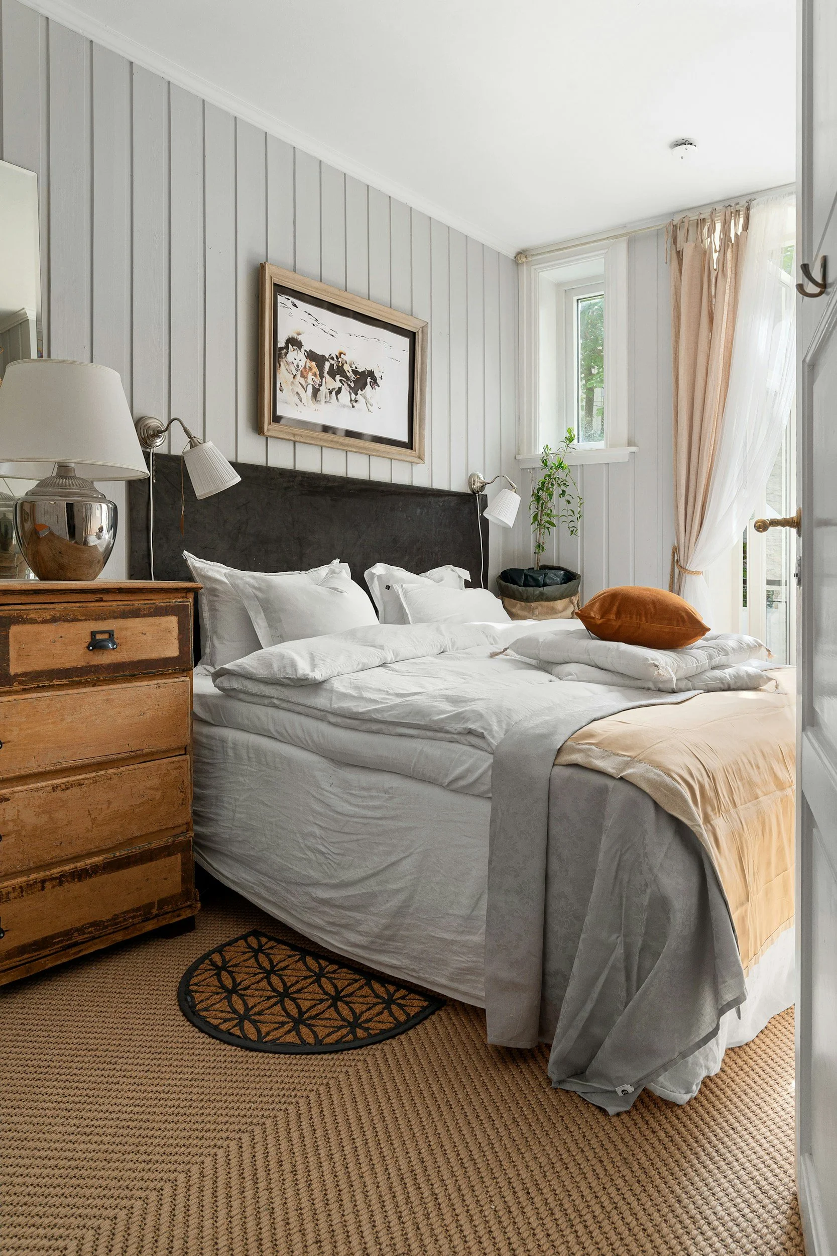 Ein Schlafzimmer mit einem unordentlichen Bett, weißen Bettdecken, sowohl grauen als auch weißen Kissen, einem braunen Kissen, einem braunen unteren Bettbezug, einem Nachttisch aus Holz, einer Deckenlampe, zwei Wandlampen über dem Bett, einem Gemälde mit Hunden, einem Fenster mit weißen Vorhängen und einer Tür zur Terrasse, die bei Tageslicht geöffnet ist.