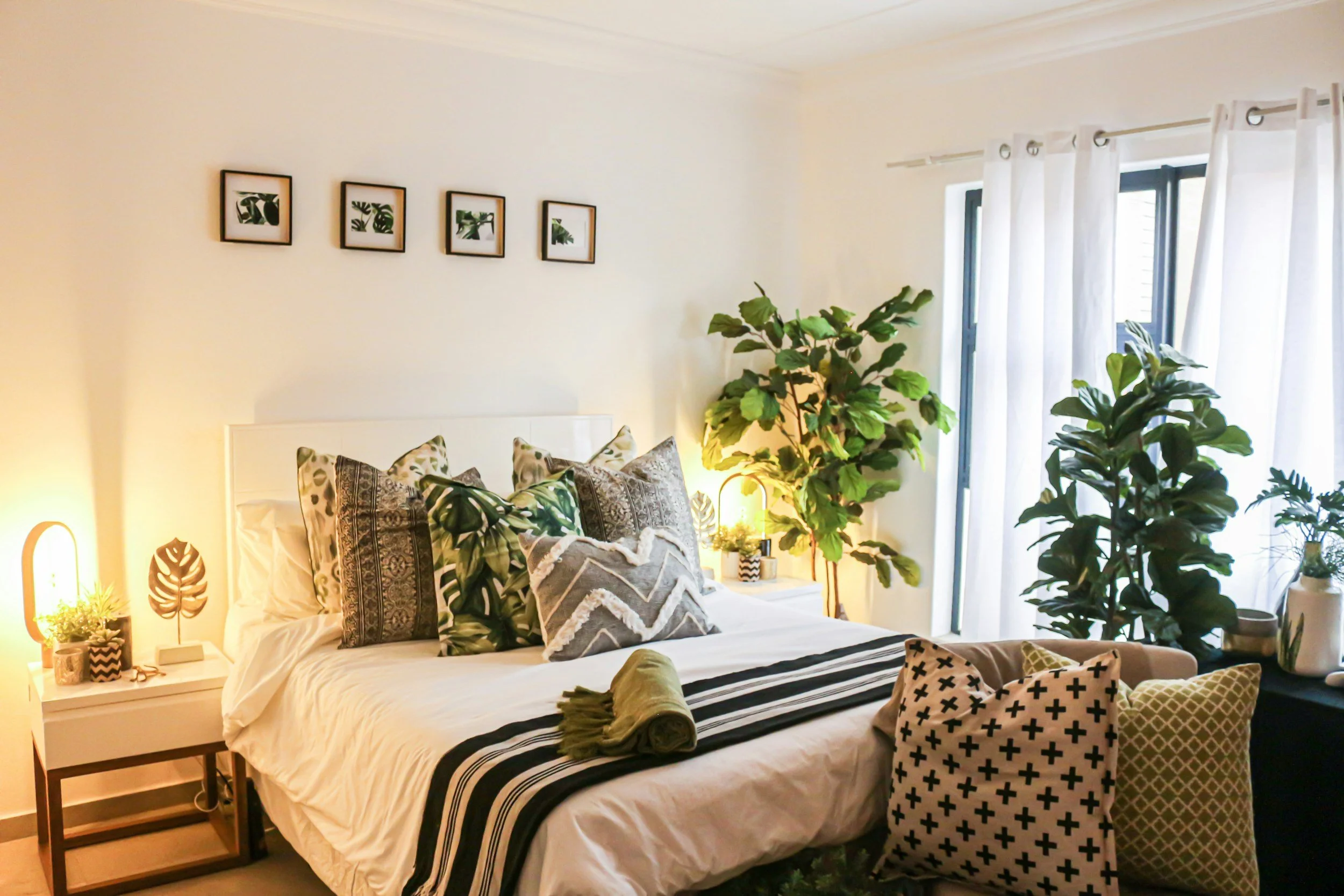 Ein Schlafzimmer mit einem weißen Bett, dekoriert mit verschiedenen Kissen, neben dem Bett steht ein Nachttisch mit einer Lampe, Pflanzen und Dekorationen, große Fenster mit weißen Vorhängen lassen viel Tageslicht herein, Wände mit 4 Wandbildern mit grünen Pflanzenmotiven.