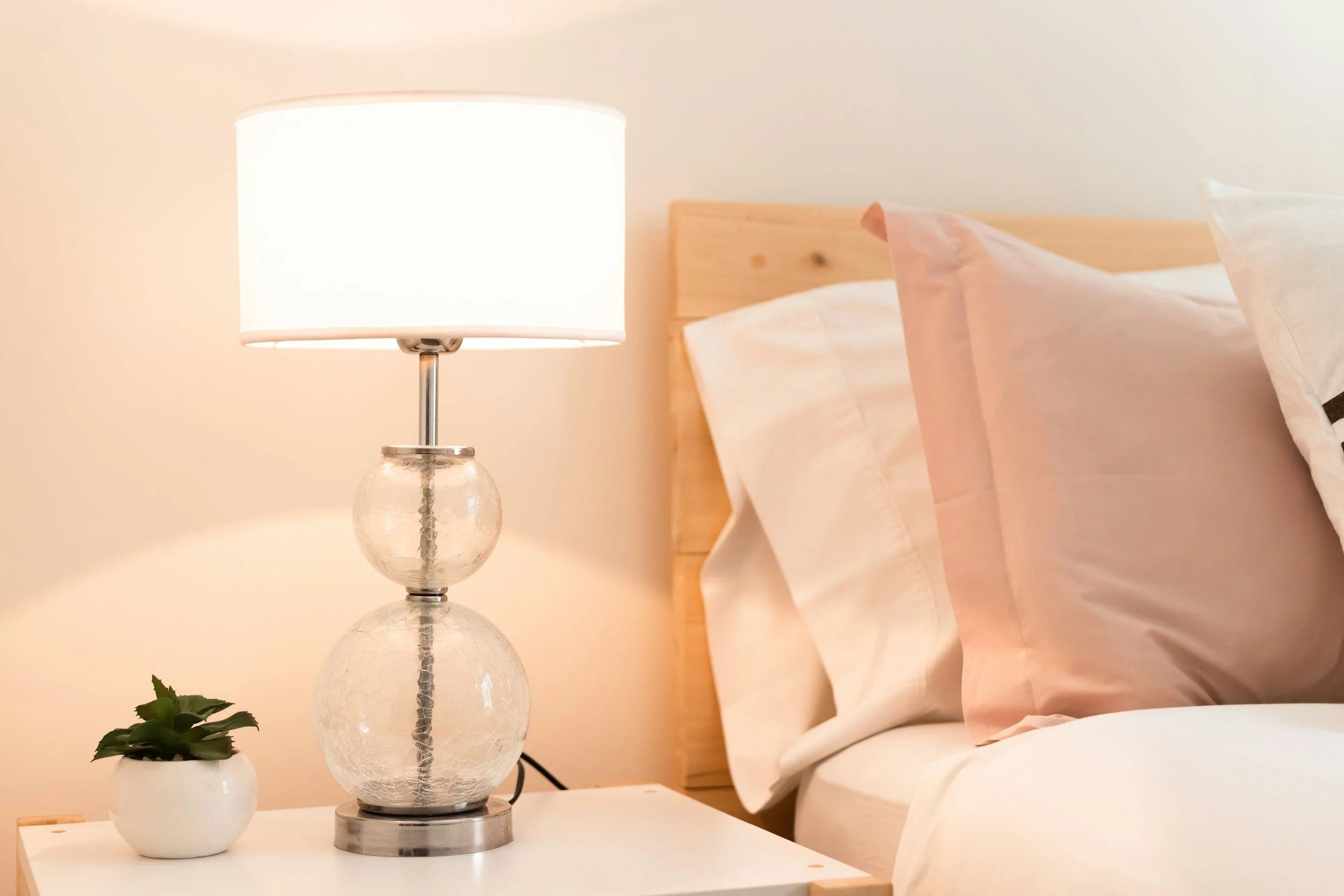 Ein Nachttisch mit einer Lampe, einer kleinen Pflanze in einer weißen Kanne, einem Kopfkissen und einem Bett mit heller Bettwäsche, Holzkopfteil, im Schlafzimmer.