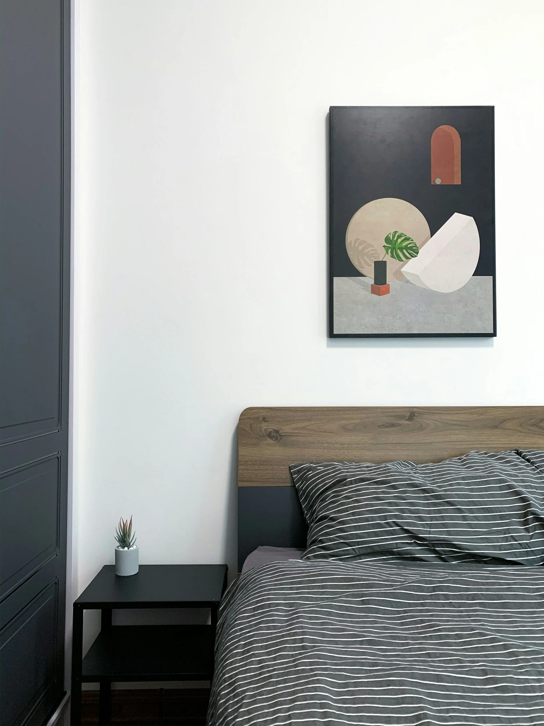 Ein Schlafzimmer mit einem Bett, das eine schwarze und weiße gestreifte Bettwäsche hat, neben einem schwarzen Nachttisch mit einer kleinen Pflanze in einer weißen Vase. Darauf hängt ein abstraktes Gemälde mit Formen und Blattsymbolen auf einem schwarzen Hintergrund.