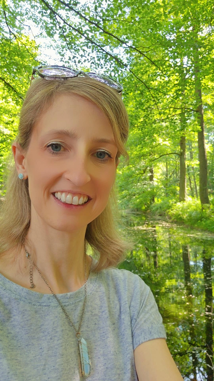 Eine Frau macht einen Selfie in einem grünen Wald mit Bäumen und einem Bach im Hintergrund.