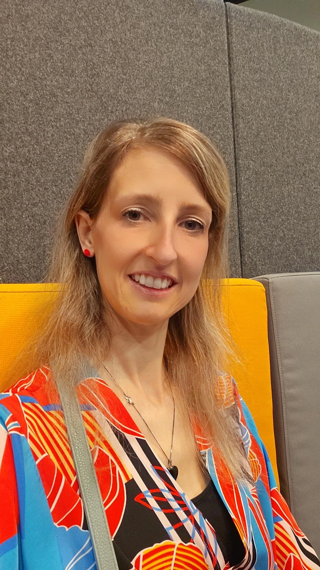 Ein Porträt einer lachenden Frau mit braunen, gewellten Haaren, die einen farbenfrohen, gemusterten Rock trägt, vor einem grauen, gepolsterten Hintergrund.