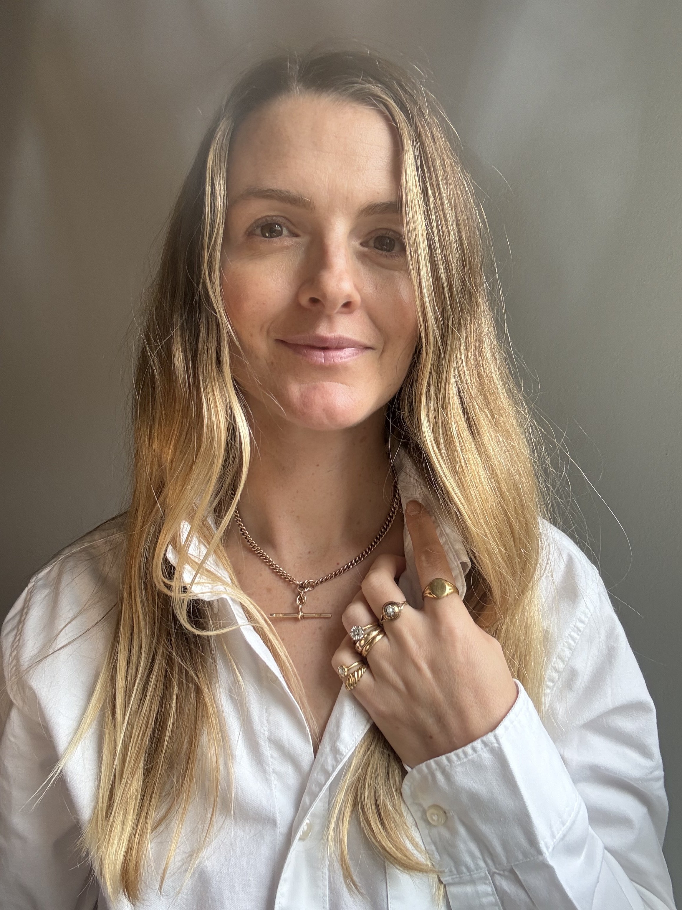 A woman with long blonde hair wearing multiple rings and necklaces, dressed in a white shirt, is posing against a plain gray background.