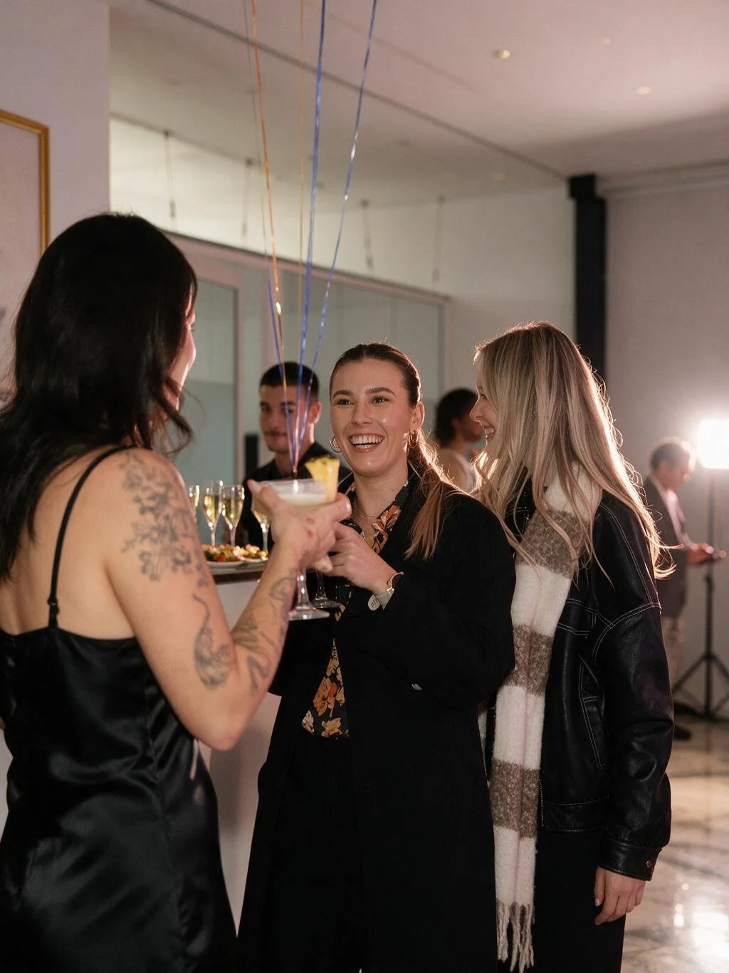Group of women smiling and chatting at a party, woman in black dress holding a cocktail with balloons attached, others with drinks, background with more guests and lighting.
