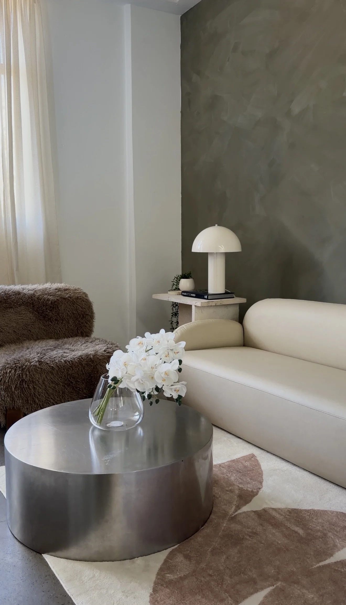 A living room with a white sofa, a brown furry armchair, a metallic round coffee table with a vase of white orchids, a small white side table with a white lamp and small potted plant, beige and brown area rugs, beige curtains, and a textured gray accent wall.