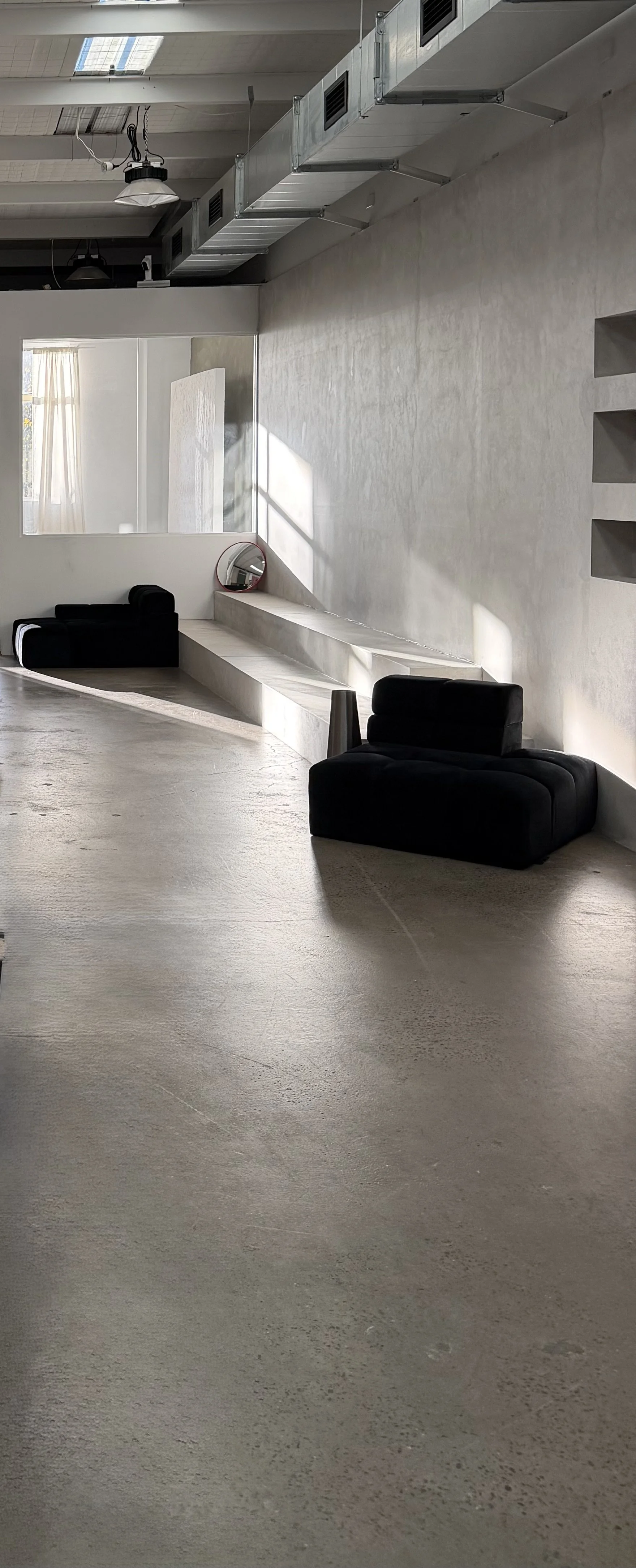 Minimalist interior space with a concrete floor, black seating, a circular mirror, and a large window with curtains letting in natural light.