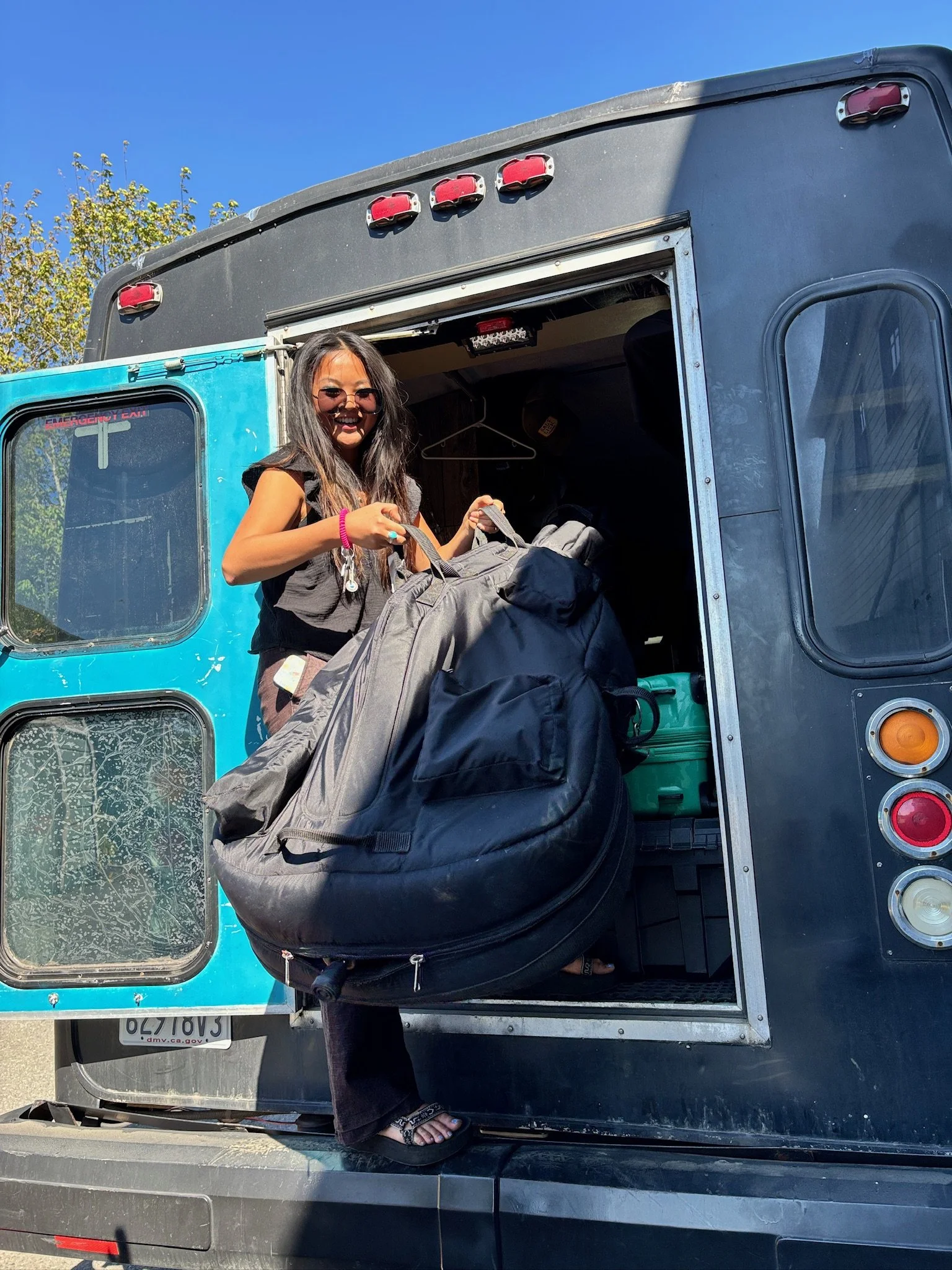 A woman with long dark hair, wearing sunglasses, a black sleeveless top, and black sandals, is standing at the open back of a food truck or bus, holding a large gray backpack. The truck has a blue and black exterior, with some greenery and a clear bl