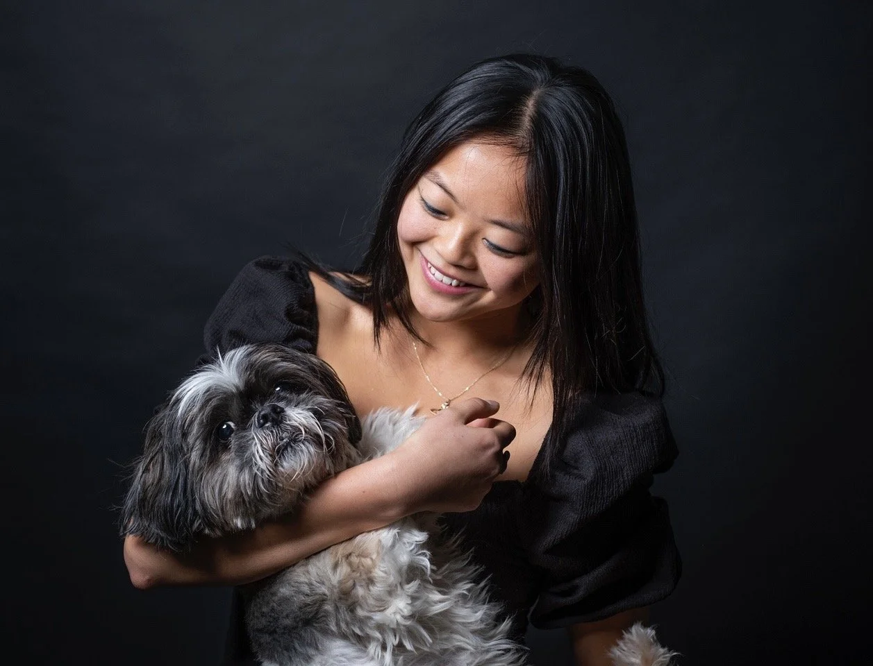 A woman holding a small dog while smiling down at it against a dark background.