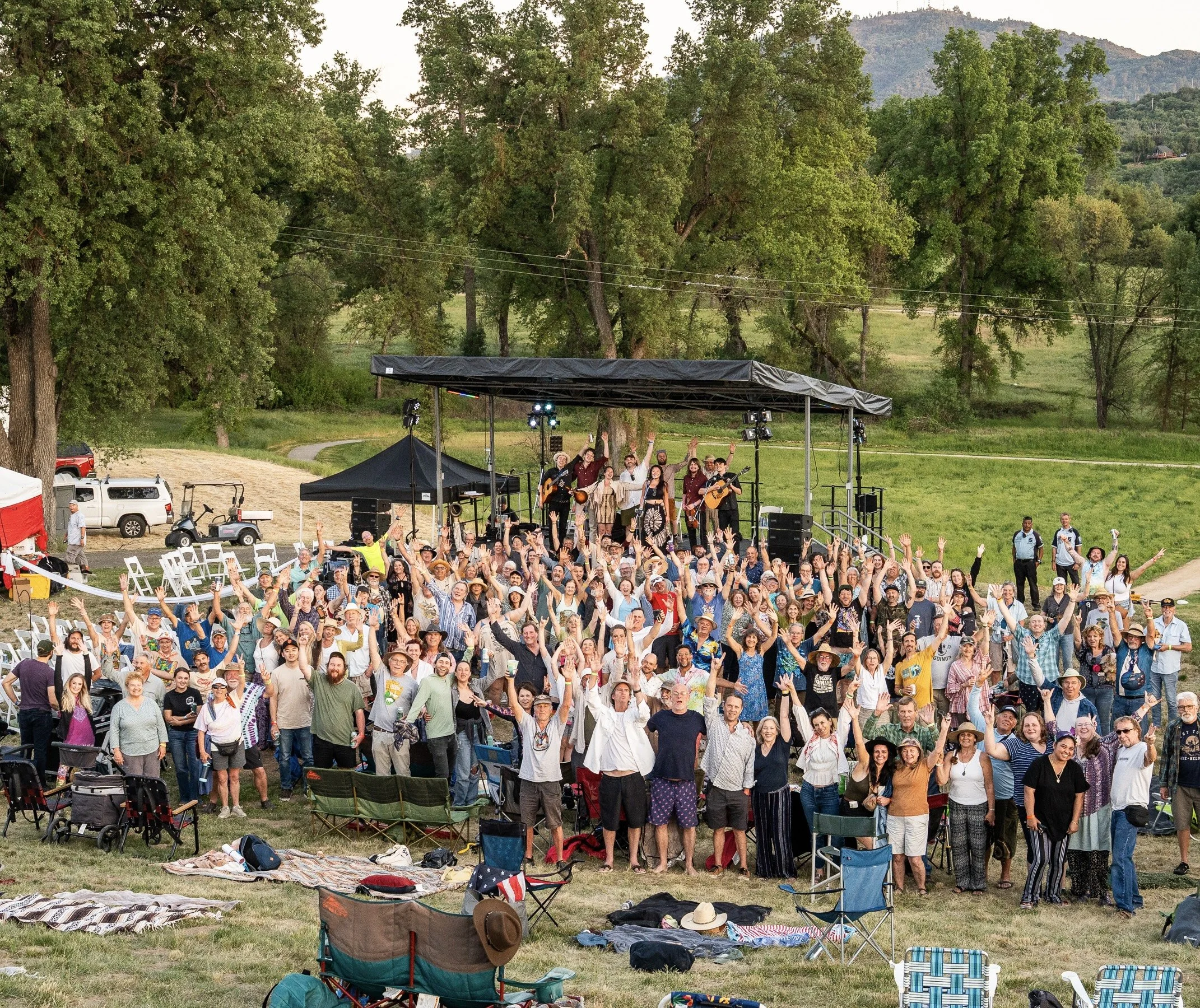 People gathered outdoors at a concert, standing and waving their hands, with musicians performing on a stage surrounded by trees, with blankets and chairs on the grass.