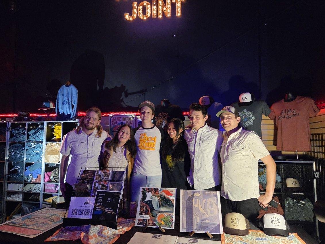 Group of people standing behind a table with merchandise at an event, with a lit sign reading 'JOIN' above them.