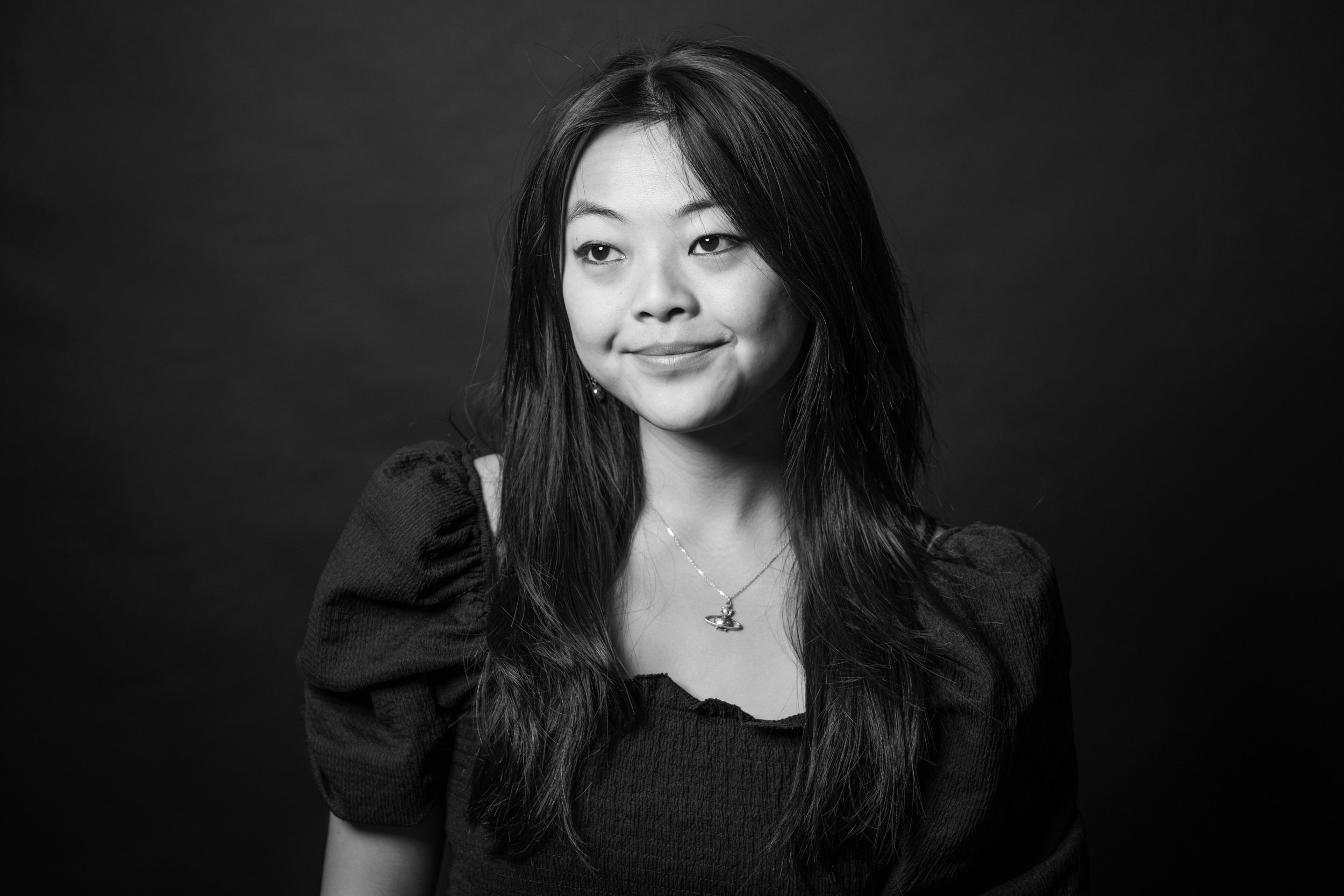 Black and white portrait of a young Asian woman with long hair, smiling softly, wearing a textured top with puffed sleeves, a necklace with a planet-themed pendant, and earrings, against a dark background.