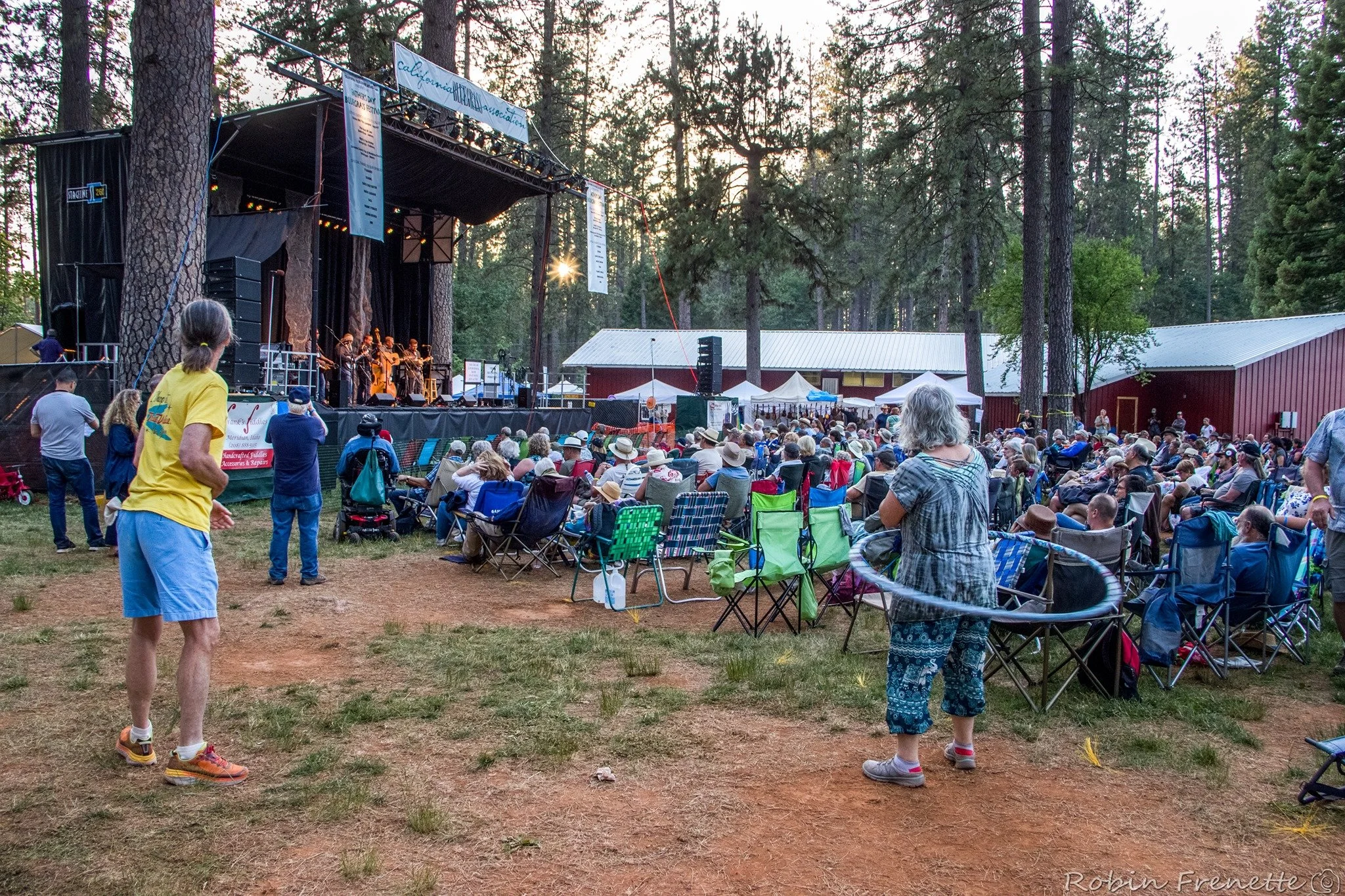 An outdoor concert in a forested area with a large crowd seated on chairs watching a band perform on stage. The audience is diverse, and many are wearing hats and casual clothing.