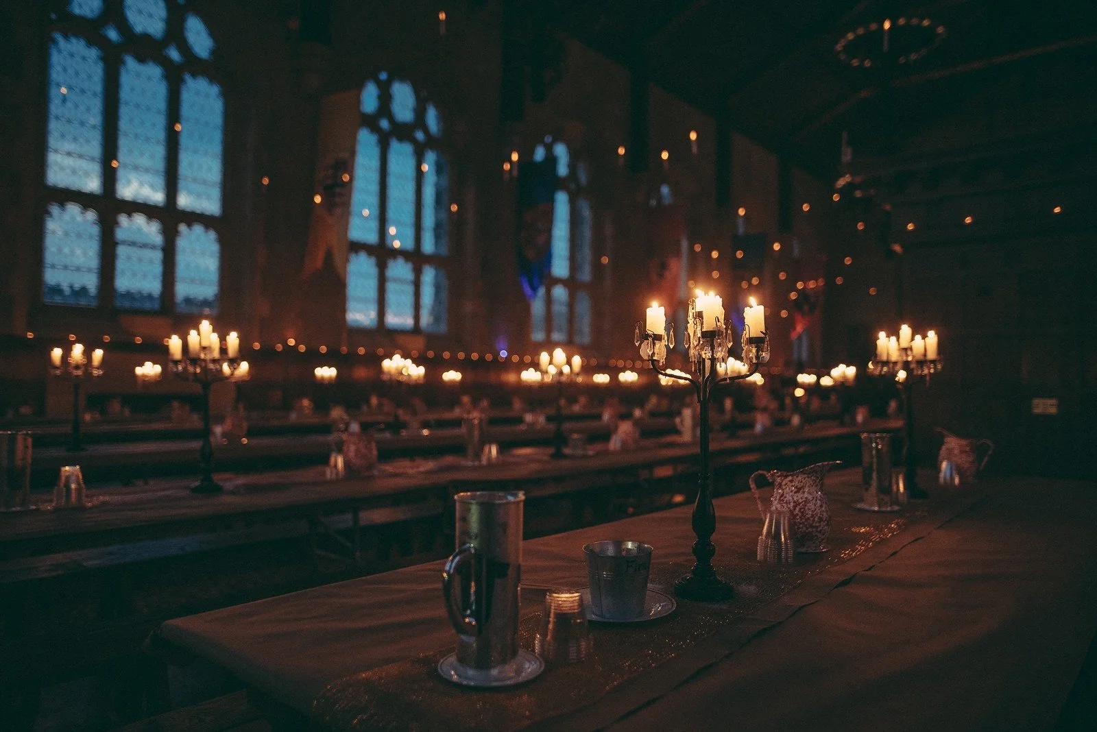 A banquet hall with stone walls and arched ceilings, decorated with artwork and statues. There are round tables with black tablecloths and white chairs, some with candles. In the foreground, there is a table with bottled water, canned drinks, snacks, and condiments. The hall appears prepared for a large gathering or event.