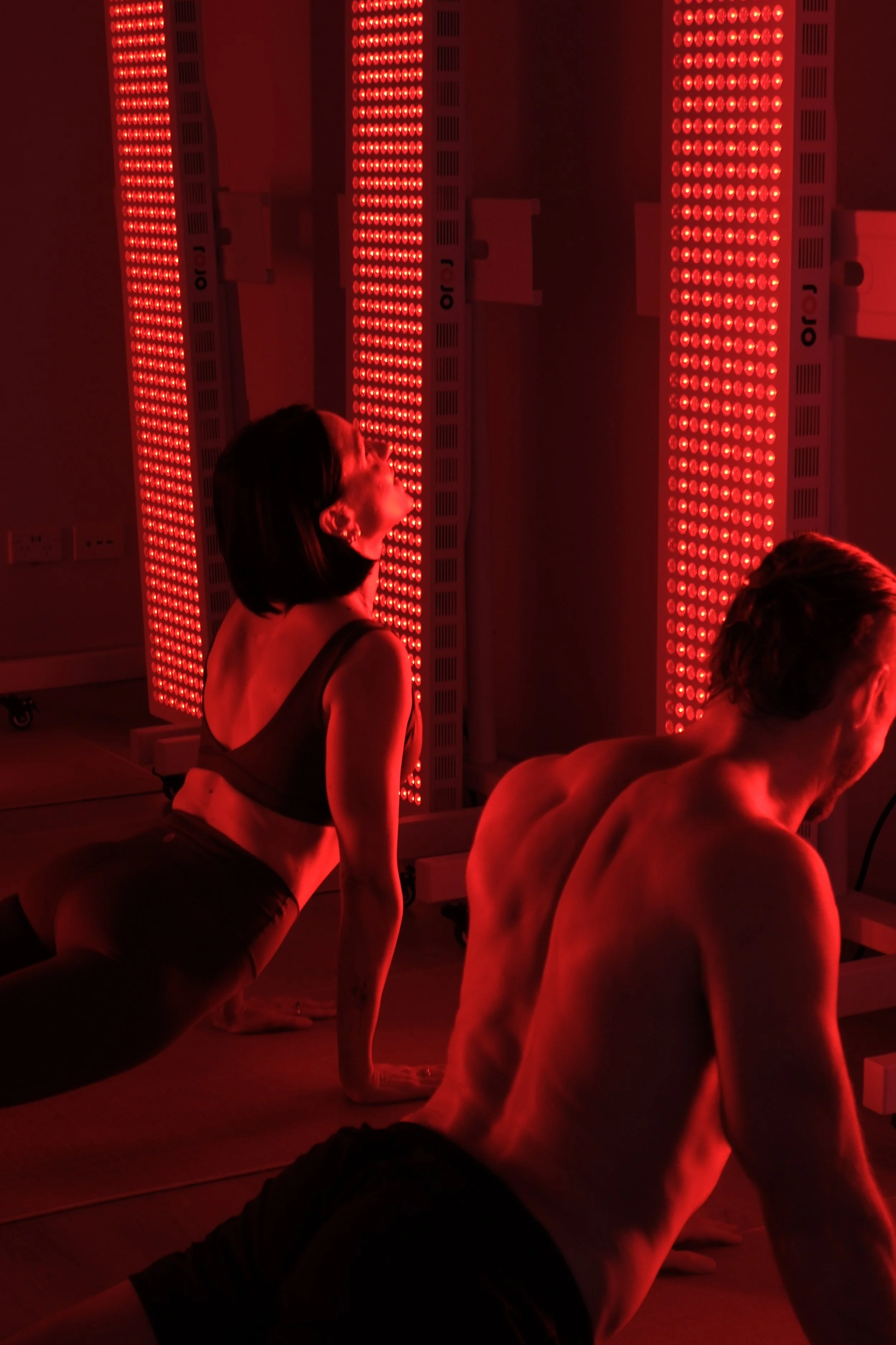 Two people practicing yoga in a room with red LED light panels.