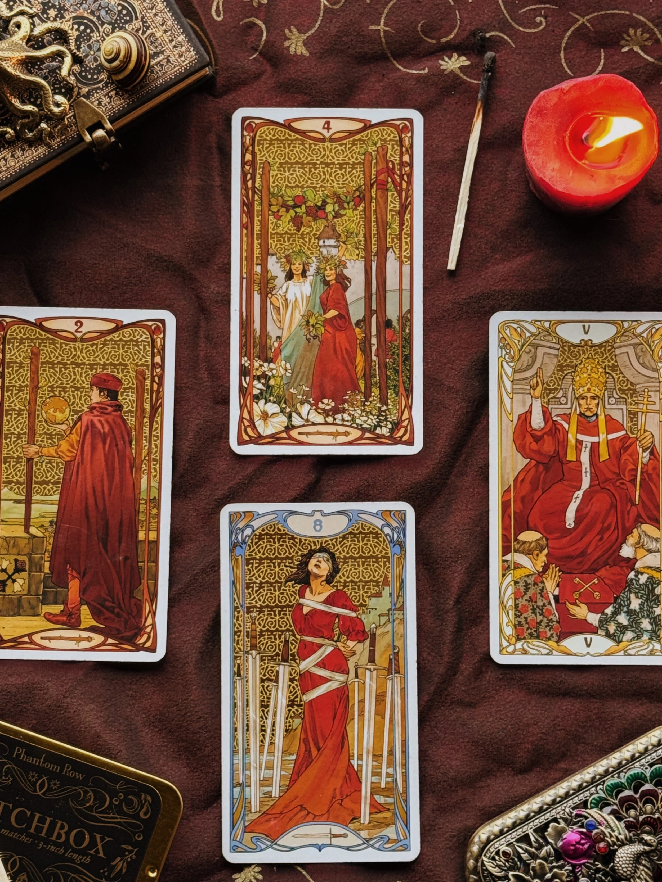 Three tarot cards laid out on a dark cloth, illuminated by three candles, with a tarot deck partially visible on the side.