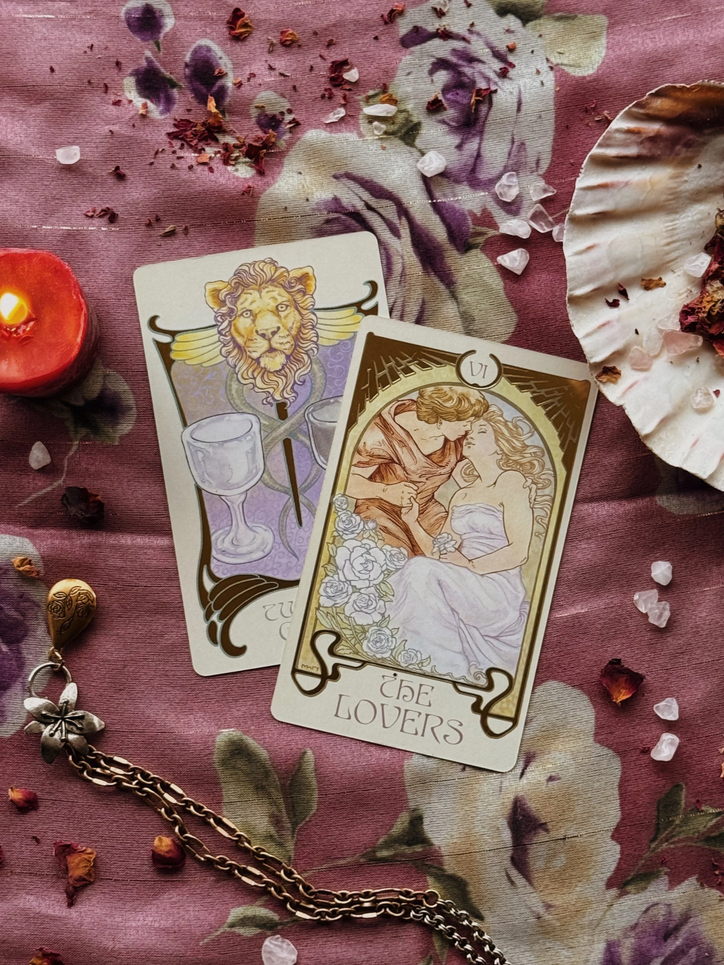 Two tarot cards, one labeled "2 of Cups" featuring a lion with wings and two drinking glasses, and the other labeled "The Lovers" showing a romantic scene of a man and woman in an embrace. The cards are on a pink fabric surface with scattered dried flower petals and a decorative shell with dried flower remnants inside.