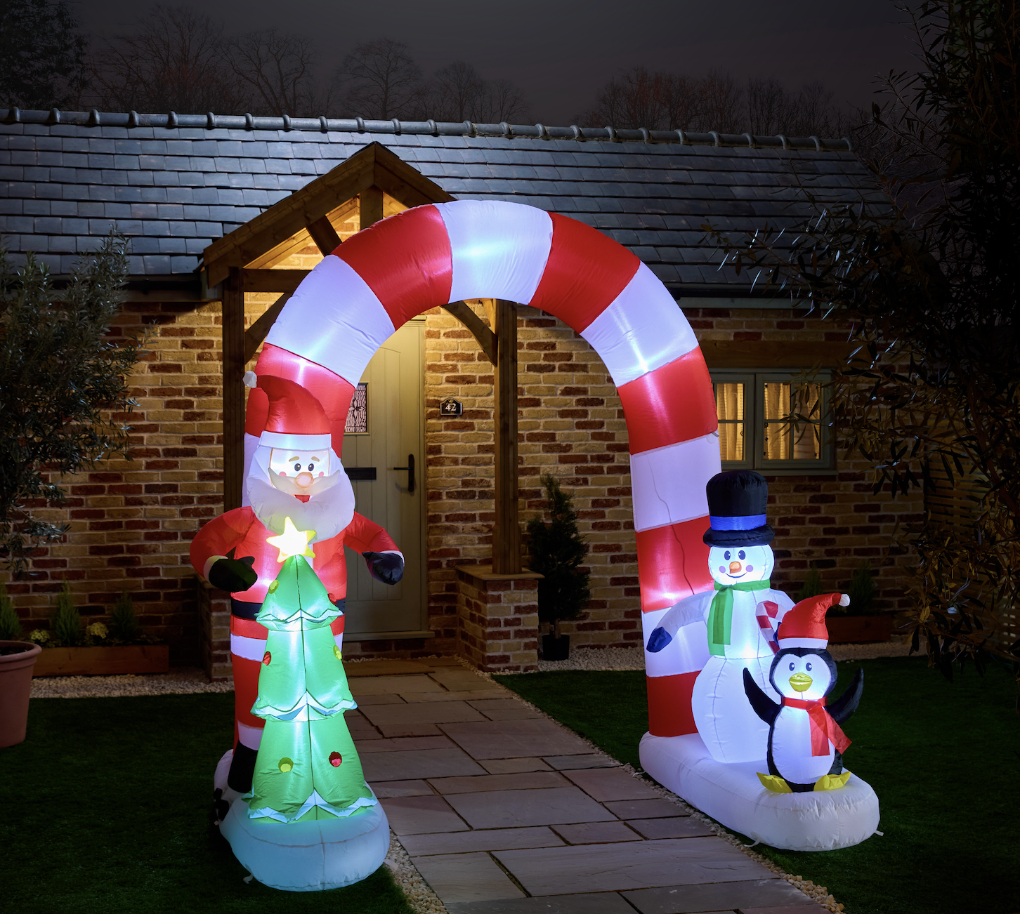 Inflatable Christmas decoration featuring Santa Claus holding a Christmas tree, a snowman wearing a top hat, and a penguin with a Santa hat, placed outside a brick house with a lit doorway, during the night.
