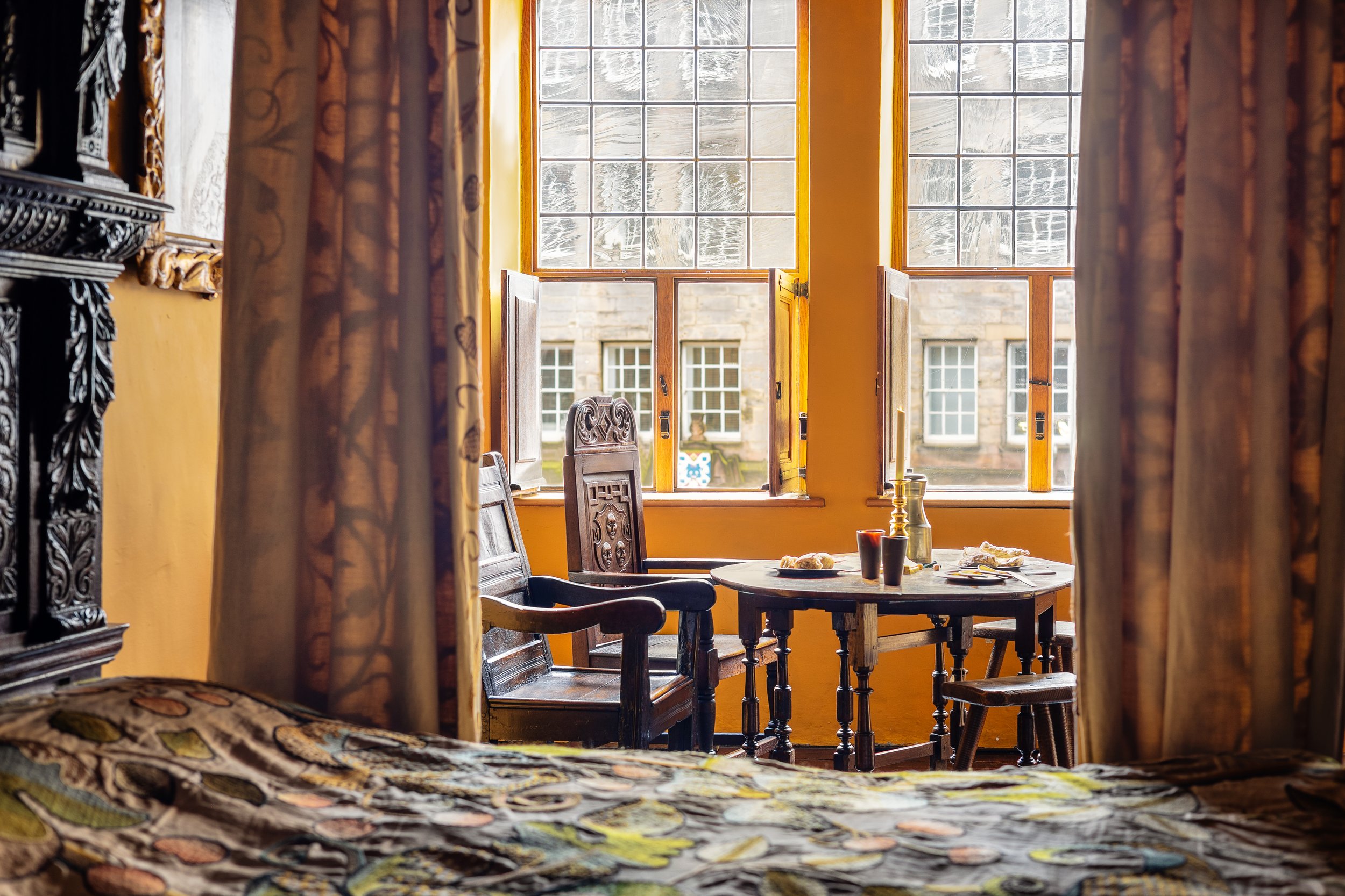 Cozy room with antique furniture, a round table set for breakfast, large patterned curtains, and an ornately carved chair near a sunlit window. Gladstone's Land, Edinburgh.