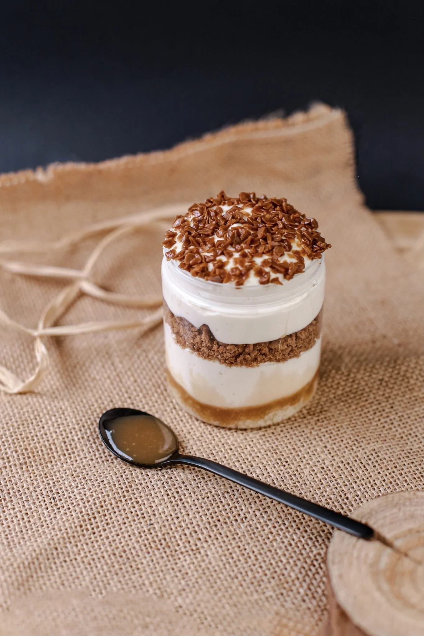 Layered dessert in a glass jar with whipped cream, chocolate shavings, cookie crumbs, and caramel sauce on top, placed on a burlap cloth with a black spoon nearby.