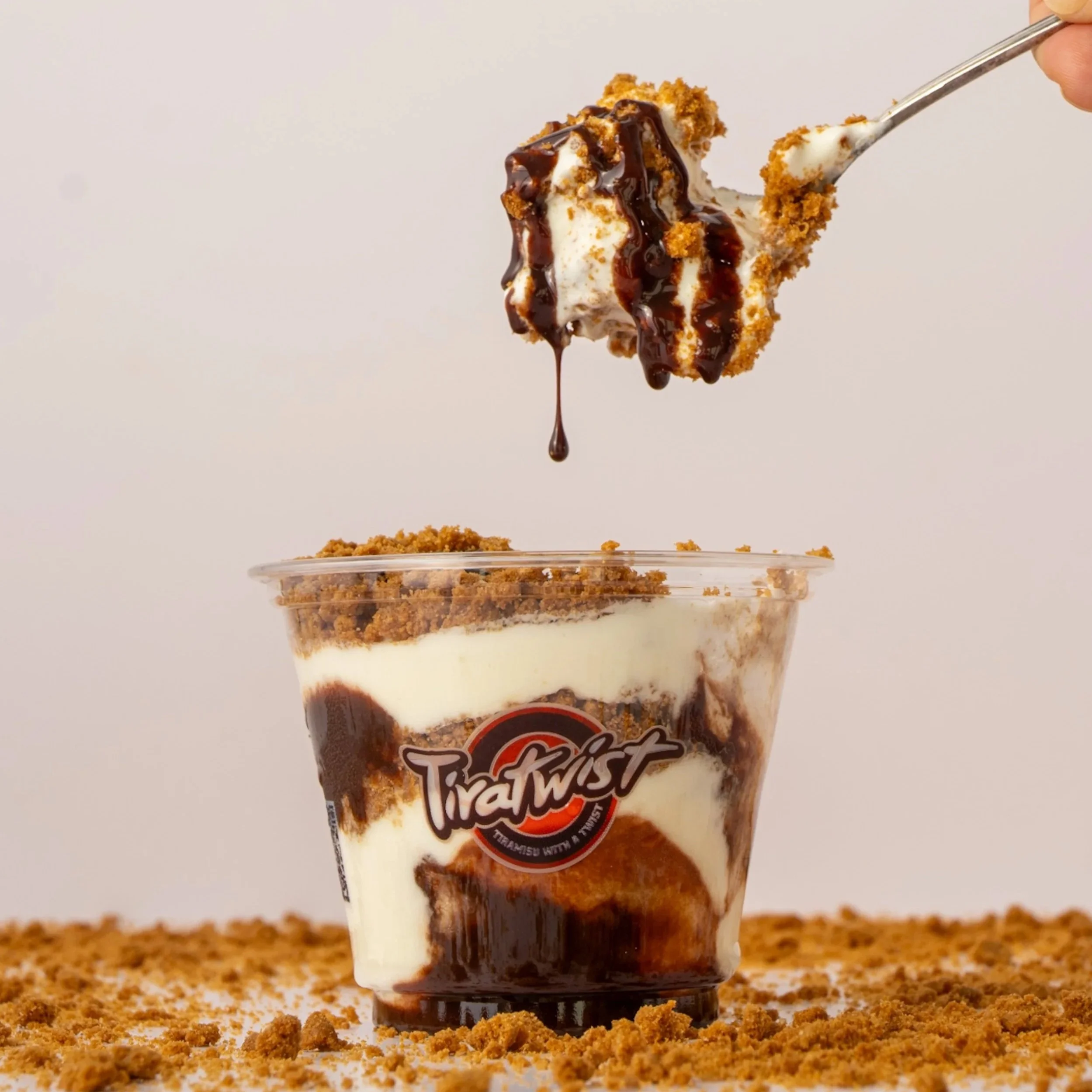 Close-up of a spoonful of layered dessert with crushed cookies, chocolate syrup, and mascarpone, being lifted above a clear cup with similar layered dessert, branded 'Tiratwist,' on a surface sprinkled with crushed cookies.