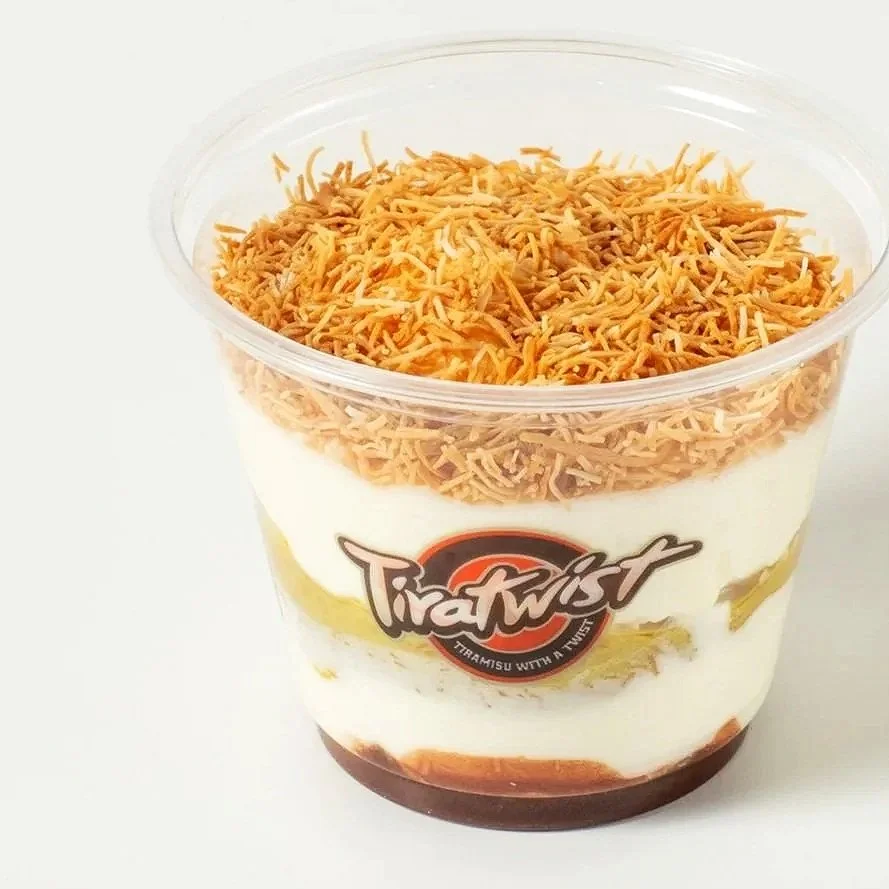 A clear plastic cup with layered dessert, topped with kataifi, branded with a 'Tira Twist' logo.