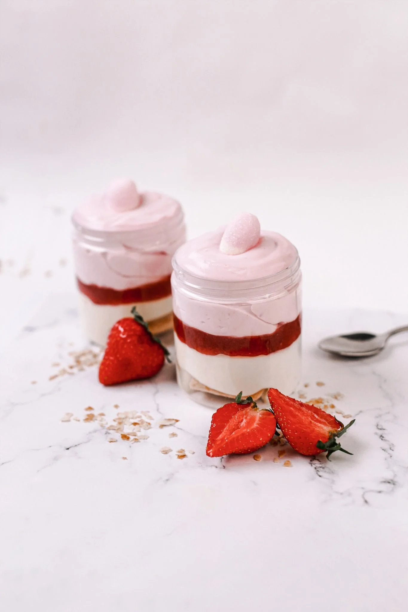 Two glass jars with layered strawberry mousse and cream, topped with pink foam and a small pink sphere, surrounded by halved strawberries and scattered oats on a white marble surface.