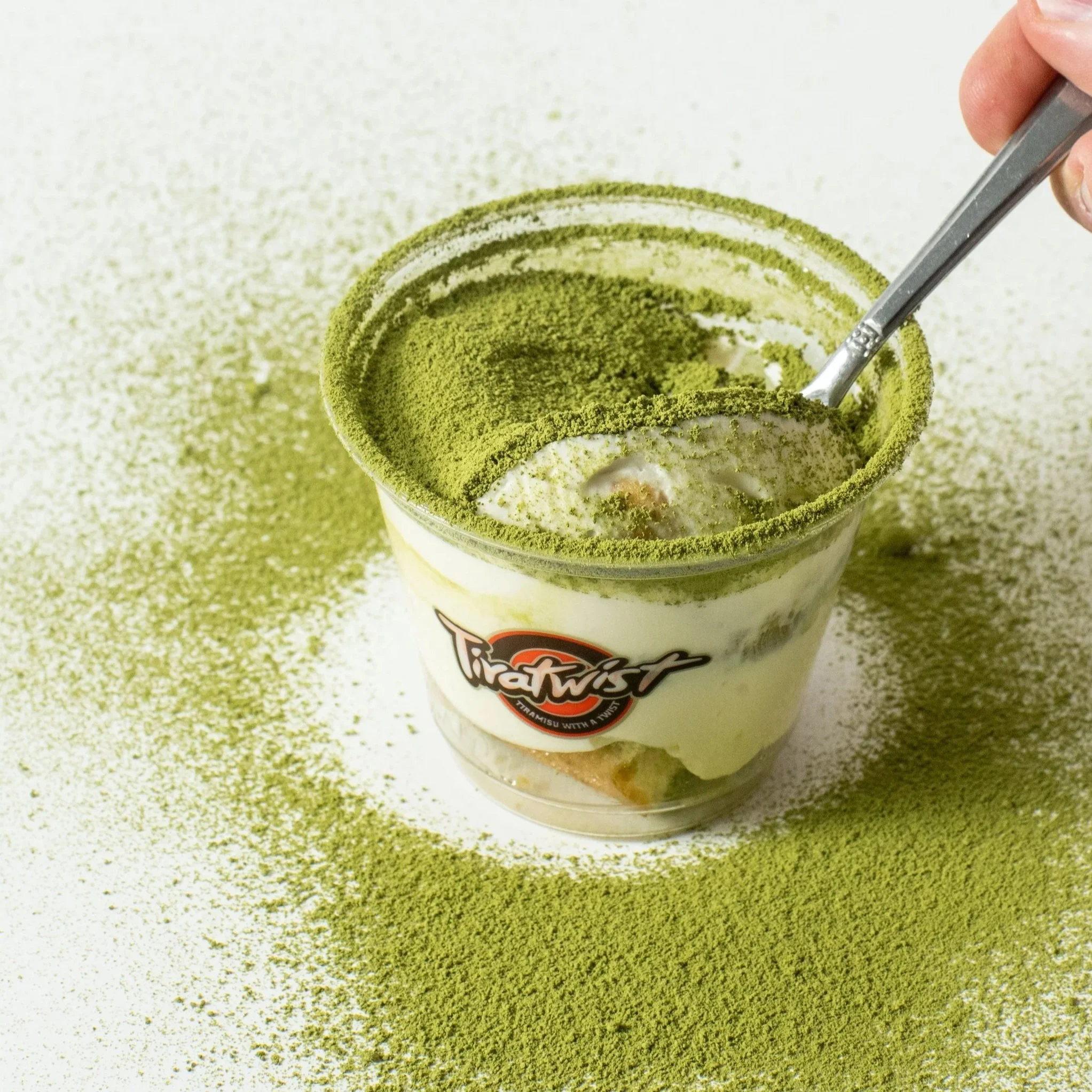 A cup of tiramisu in a plastic container sprinkled with matcha powder, with a spoon inside and some powder scattered nearby.