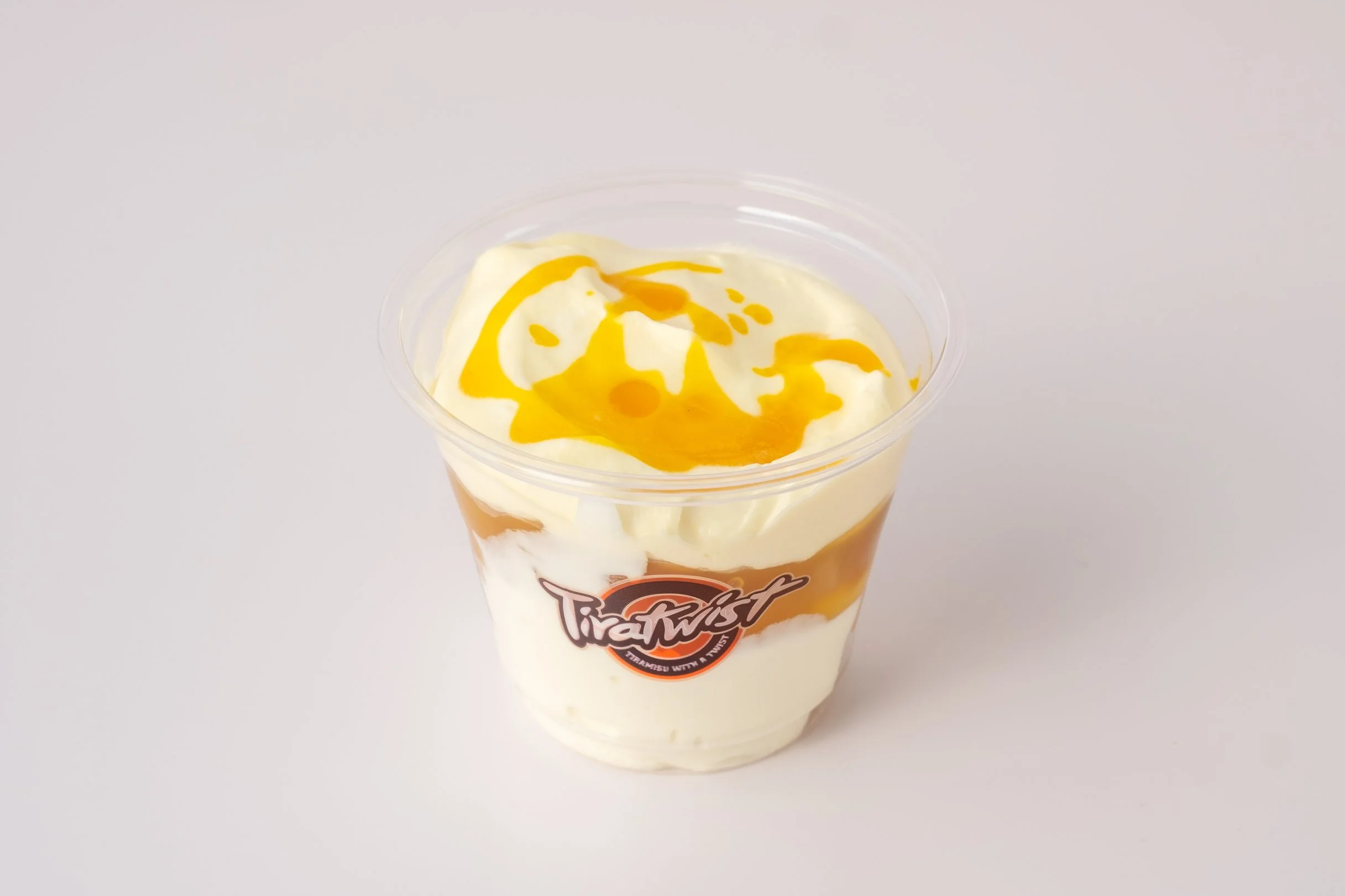 A clear plastic cup of cream with caramel sauce and yellow syrup drizzle, featuring a TiraTwist logo on the front, placed on a plain white surface.