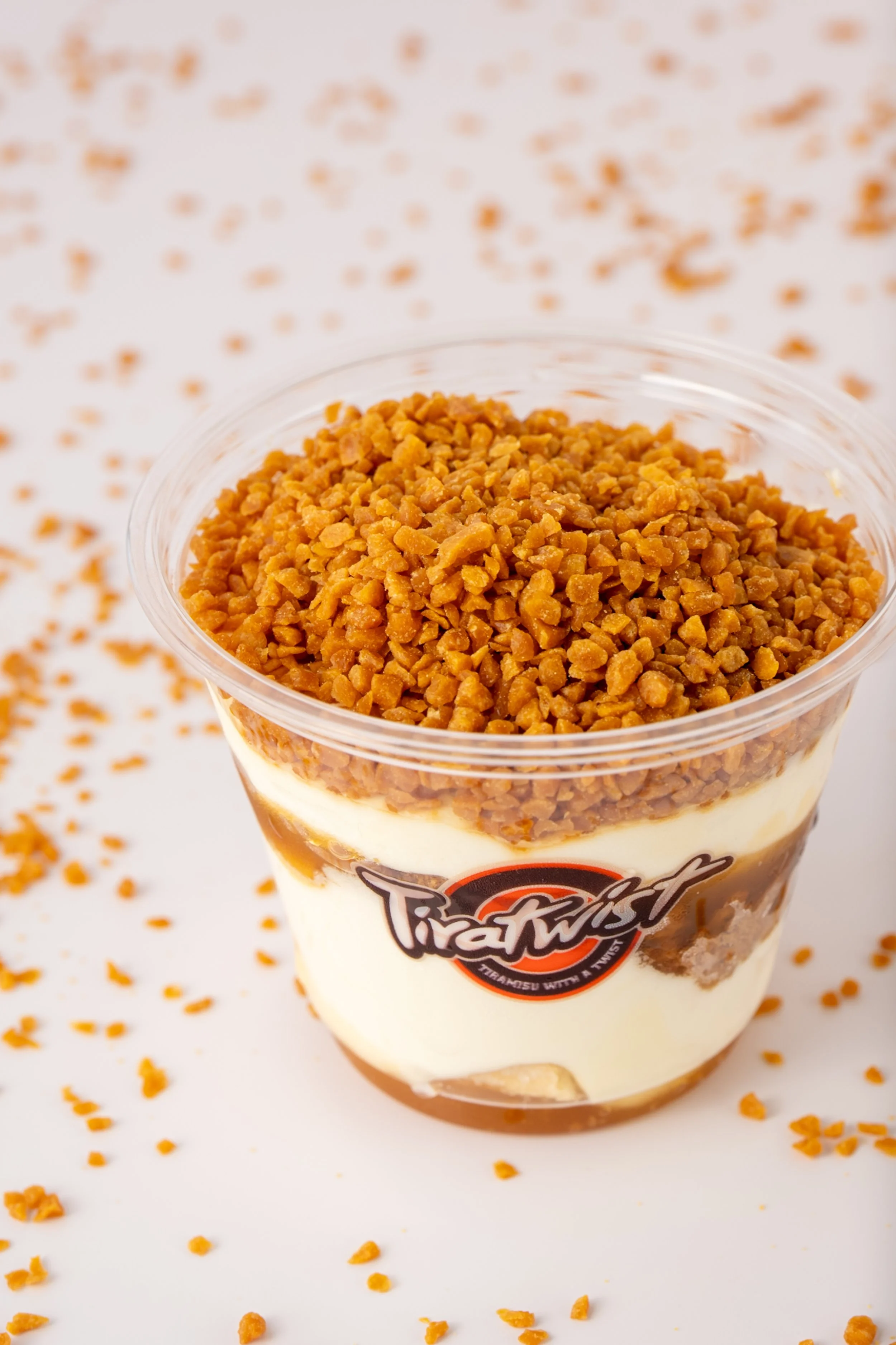 A plastic cup filled with layered tiramisu, topped with chopped caramelized pieces.