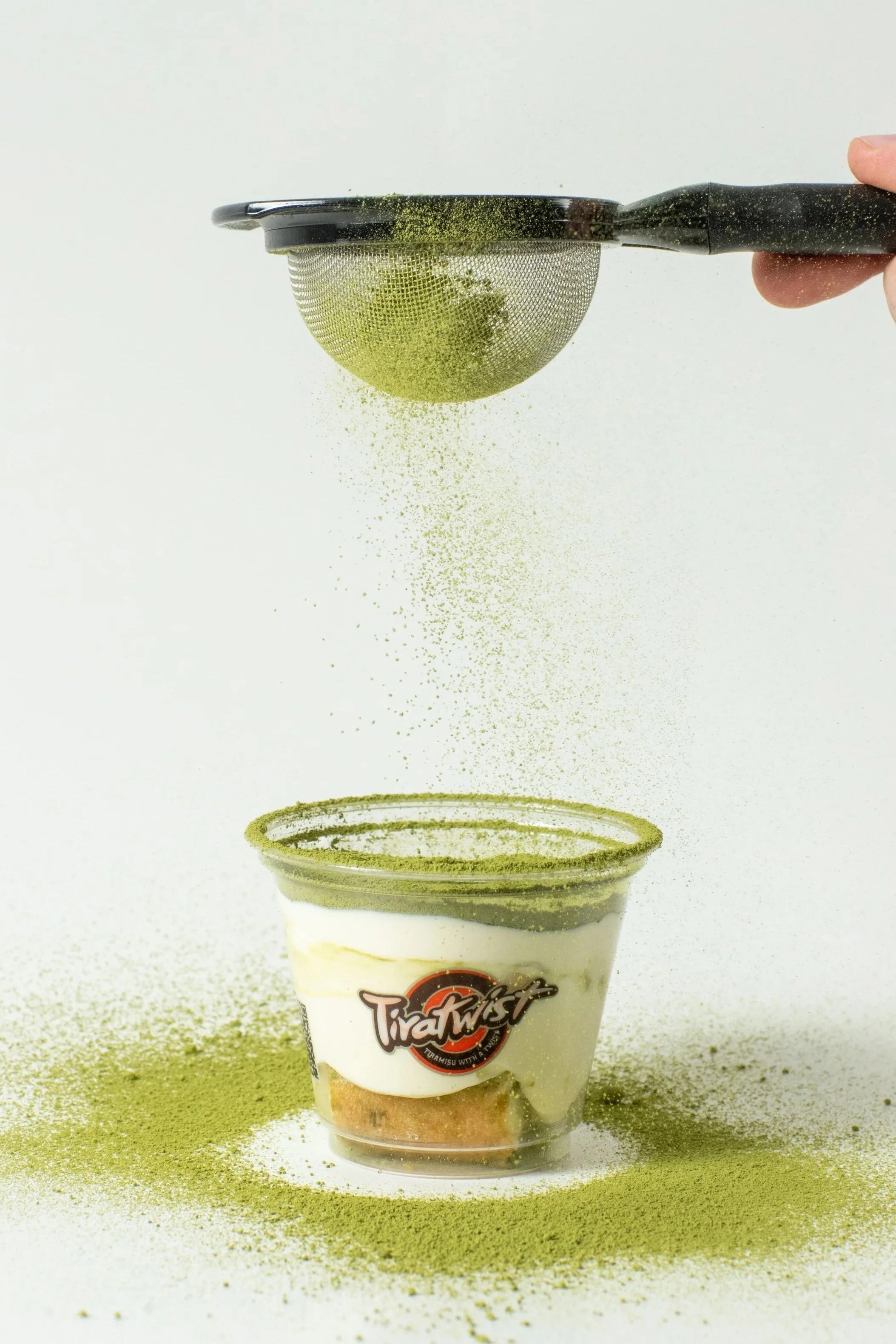 A hand holding a fine mesh strainer sprinkling green powder over a dessert cup labeled 'Tiratwist' on a white background.