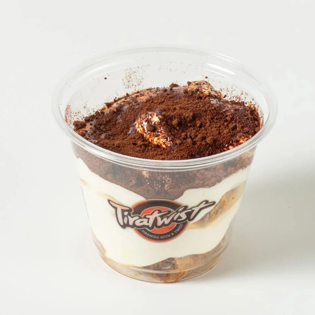 Tira Twist tiramisu dessert in a clear plastic cup with layers of cream and cocoa powder on top, placed on a white surface.