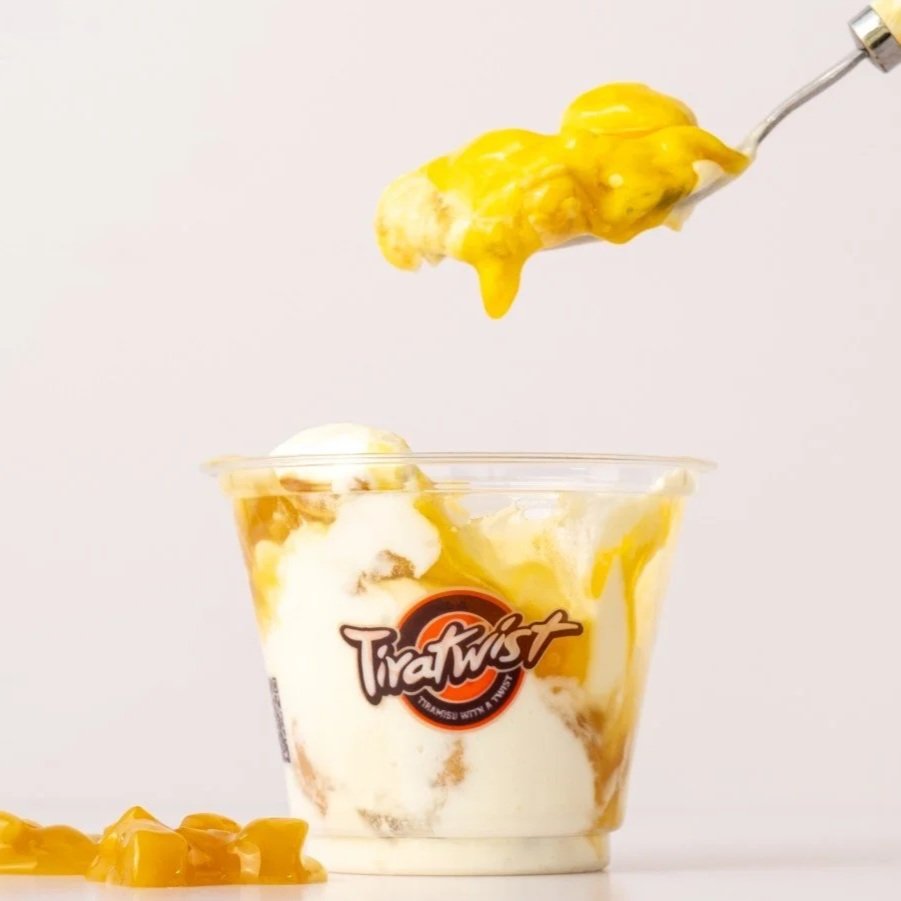 A clear plastic cup filled with cream topped with a spoonful of mango chunks and mango syrup dripping down. The cup has a TiraTwist logo on it, and some mango pieces are on the surface below.