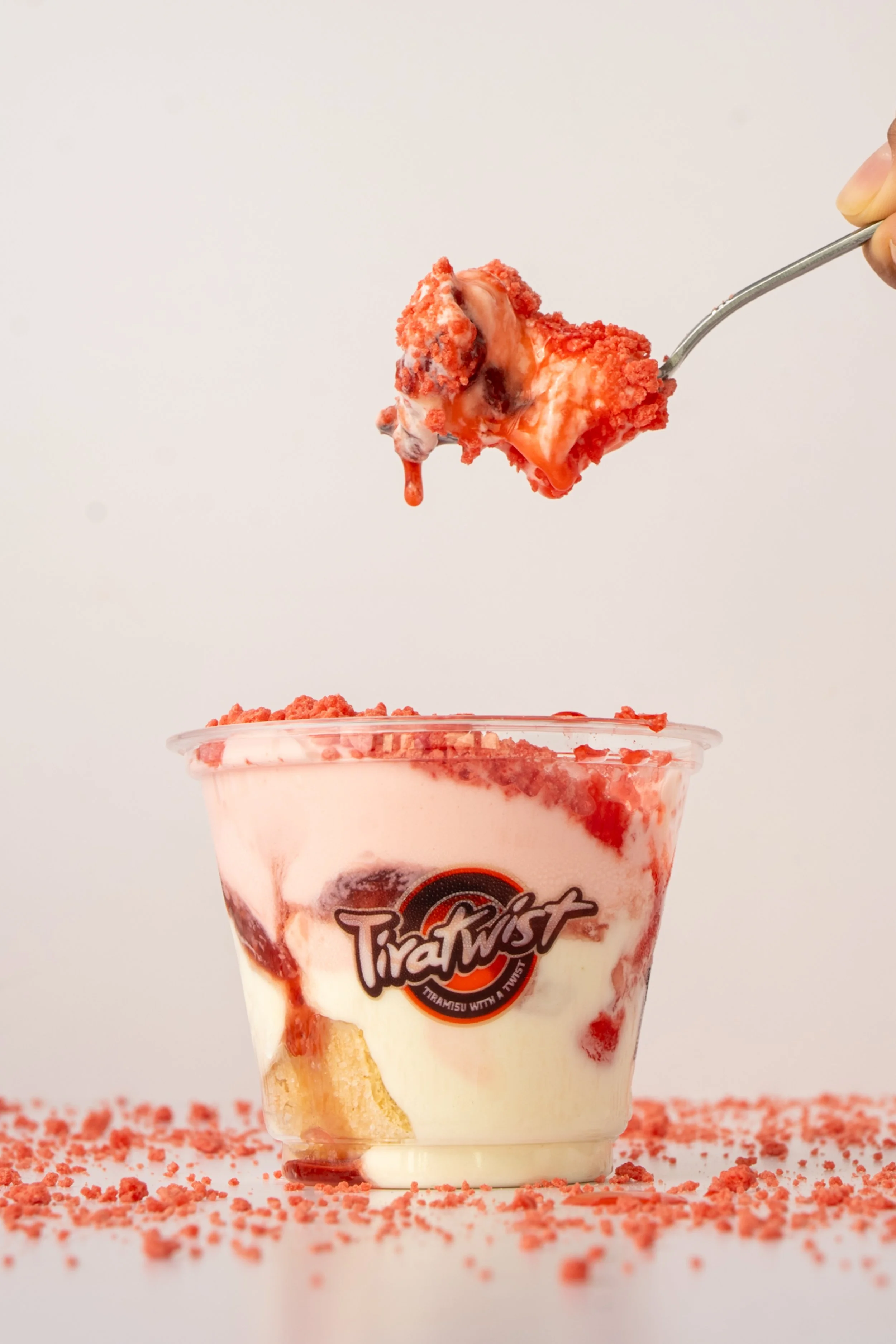 A transparent cup filled with a dessert containing cream, red fruit sauce, and crumbled cookies, with a spoon holding a piece of the dessert above the cup. The cup has a "Tira Twist" logo on it, and red crumbs are scattered around the base on a white background.