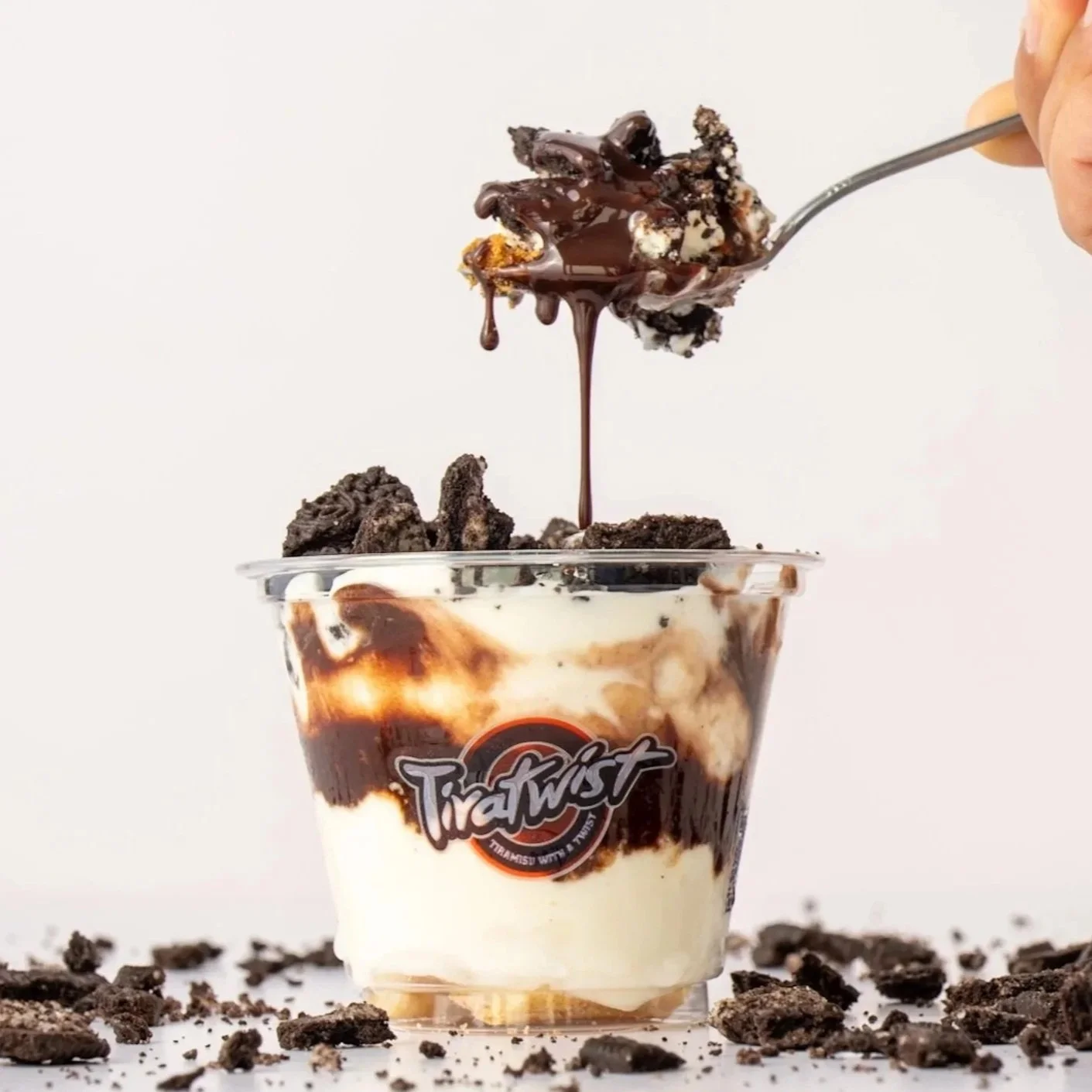 Oreo cookie rocks with a spoonful of mascarpone topped with chocolate sauce in a clear cup labeled 'Tiratwist'