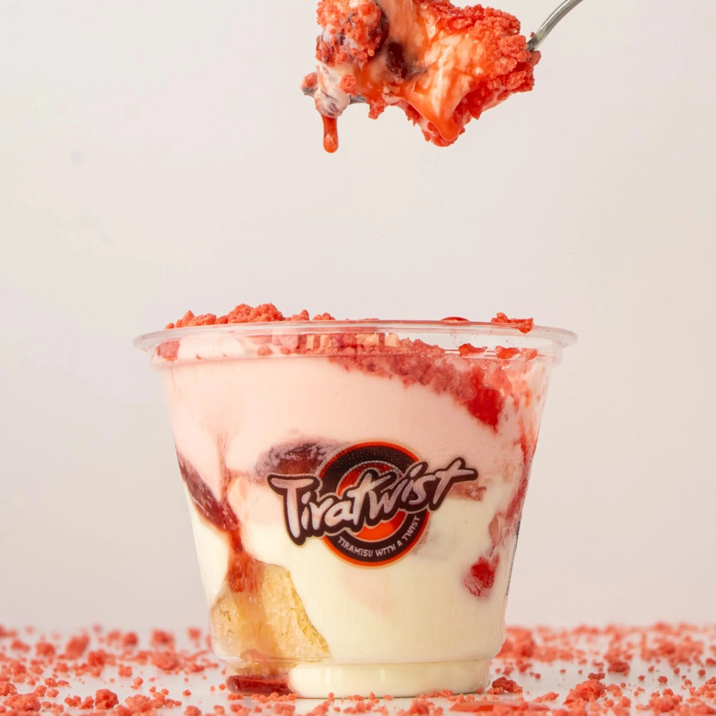 A layered dessert in a clear jar with pink frosting, a small pink candy on top, a layer of red fruit filling, white cream, and a cookie at the bottom.