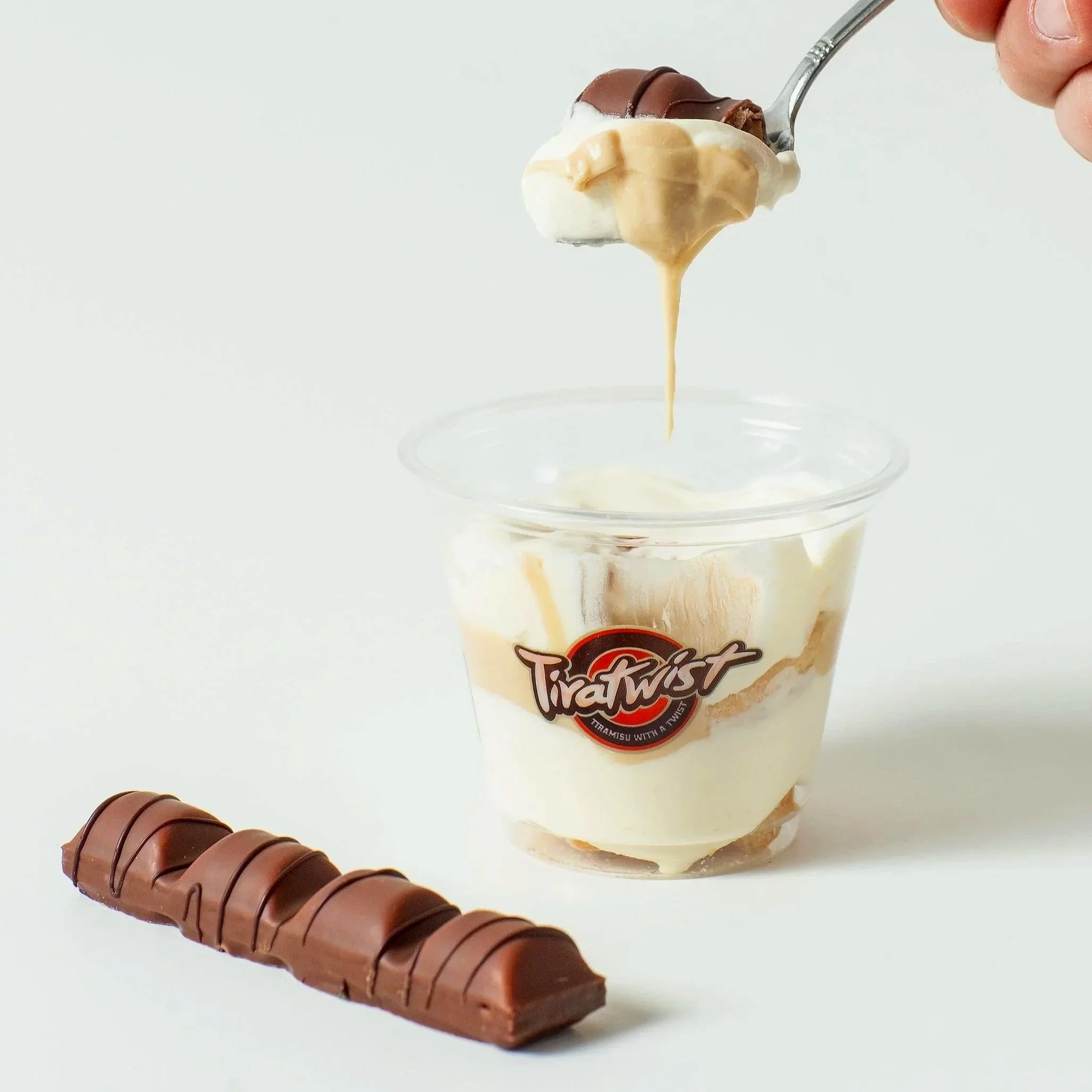 A hand holding a spoon with tiramisu and hazelnut syrup over a clear plastic cup, with a chocolate-covered wafer bar on a white surface and a white background.