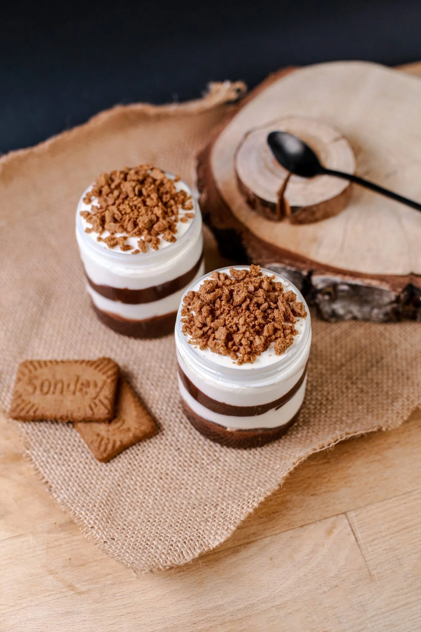 Two jars of layered dessert with whipped cream, chocolate, and crumbled cookies on a burlap cloth, with cookies and wood slice in the background.