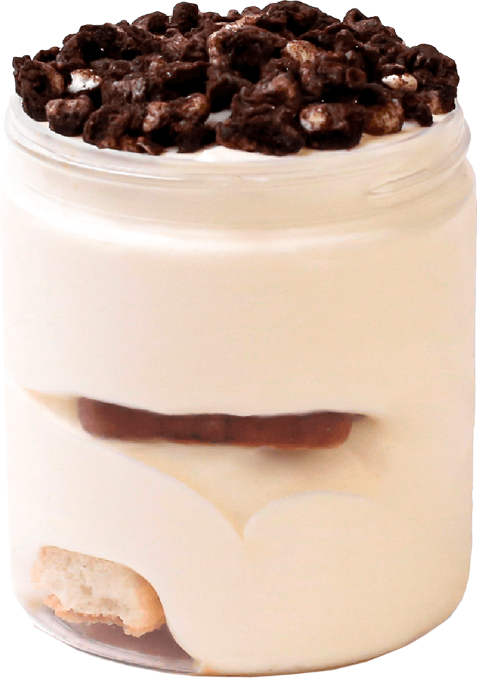 Close-up of a layered dessert in a glass jar with whipped cream, chocolate pieces, and a biscuit layer.