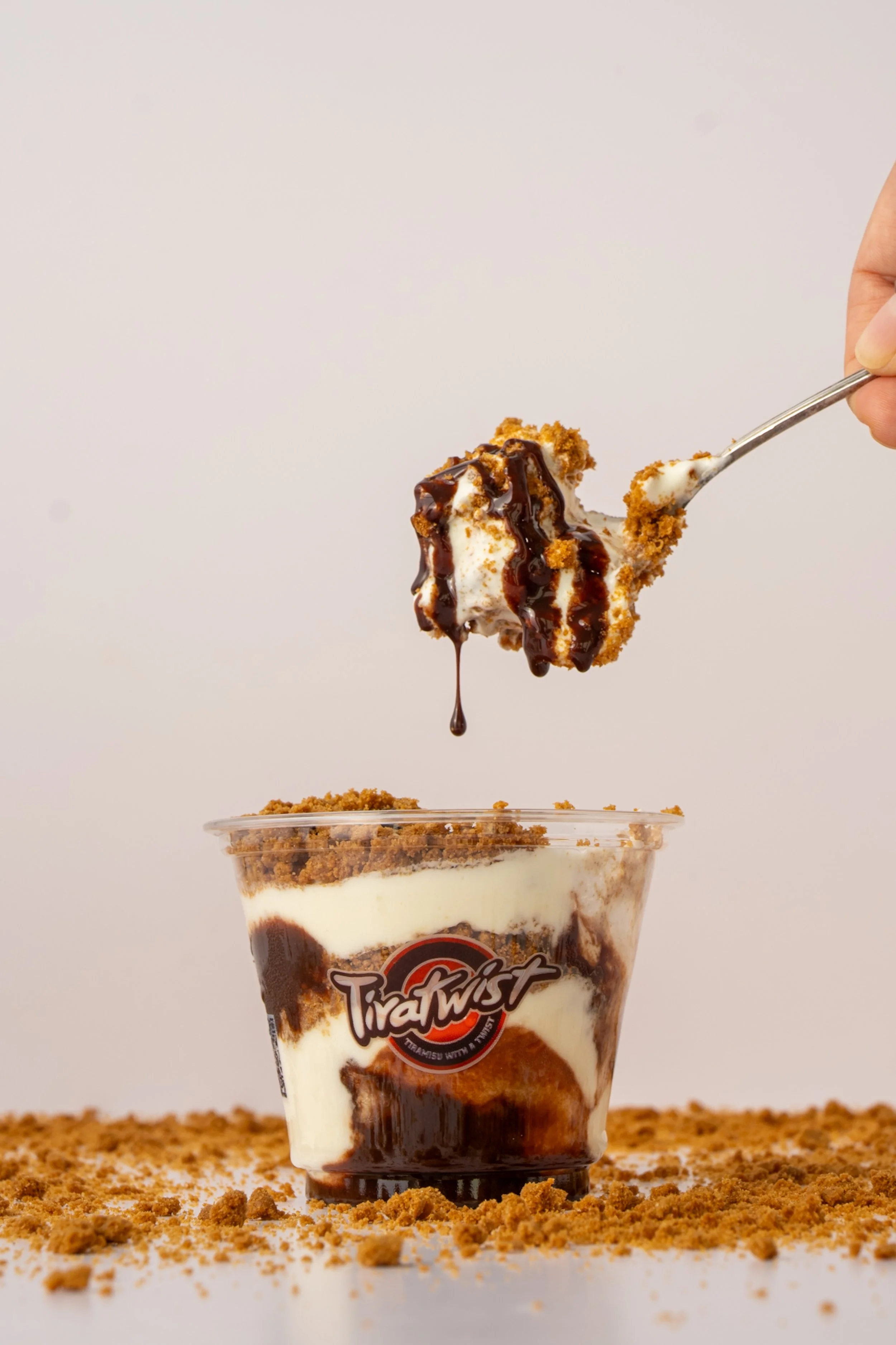 A hand holding a spoon with a scoop of cream topped with chocolate syrup over a cup of tiramisu, which is placed on a table with cookie crumbs. The cup has a "TiraTwist" logo on it.
