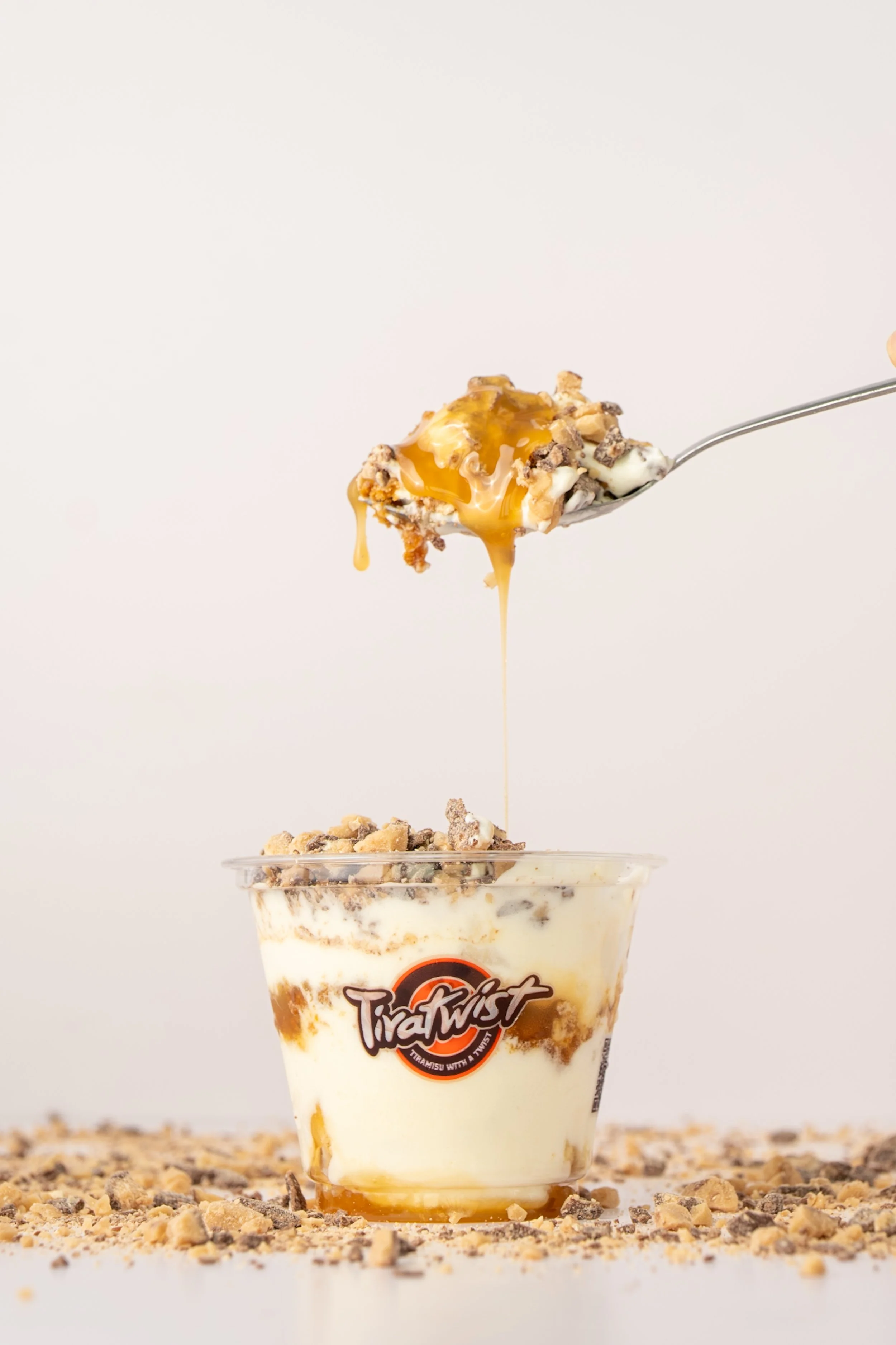 A dessert cup with caramel, toffee, and cream, topped with crushed cookies, with a spoon lifting some onto the cup.