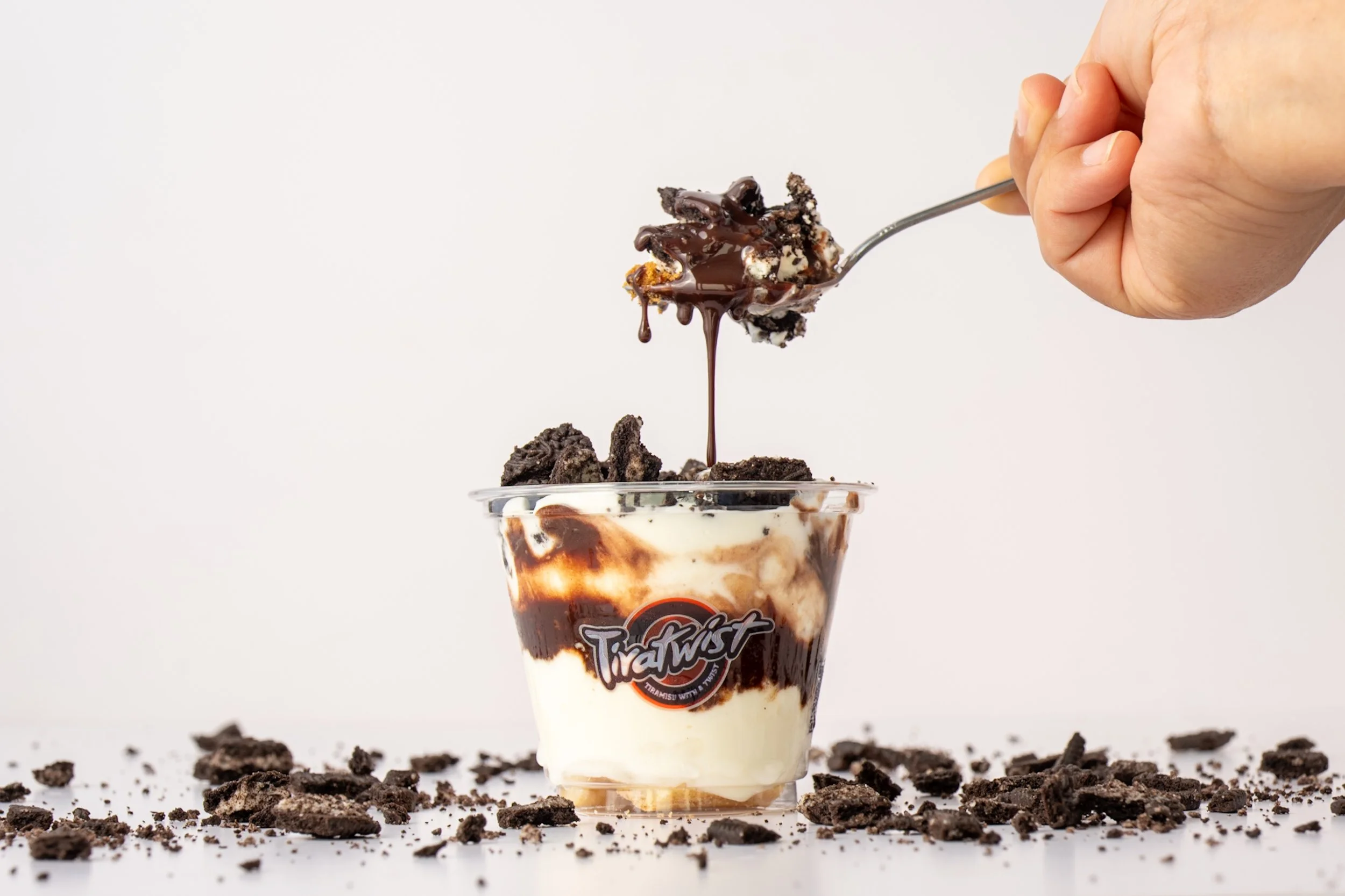 Hand holding a spoon with an Oreo cookie and chocolate sauce over a Oreo tiramisu cup, with crumbled Oreos scattered on a white surface.